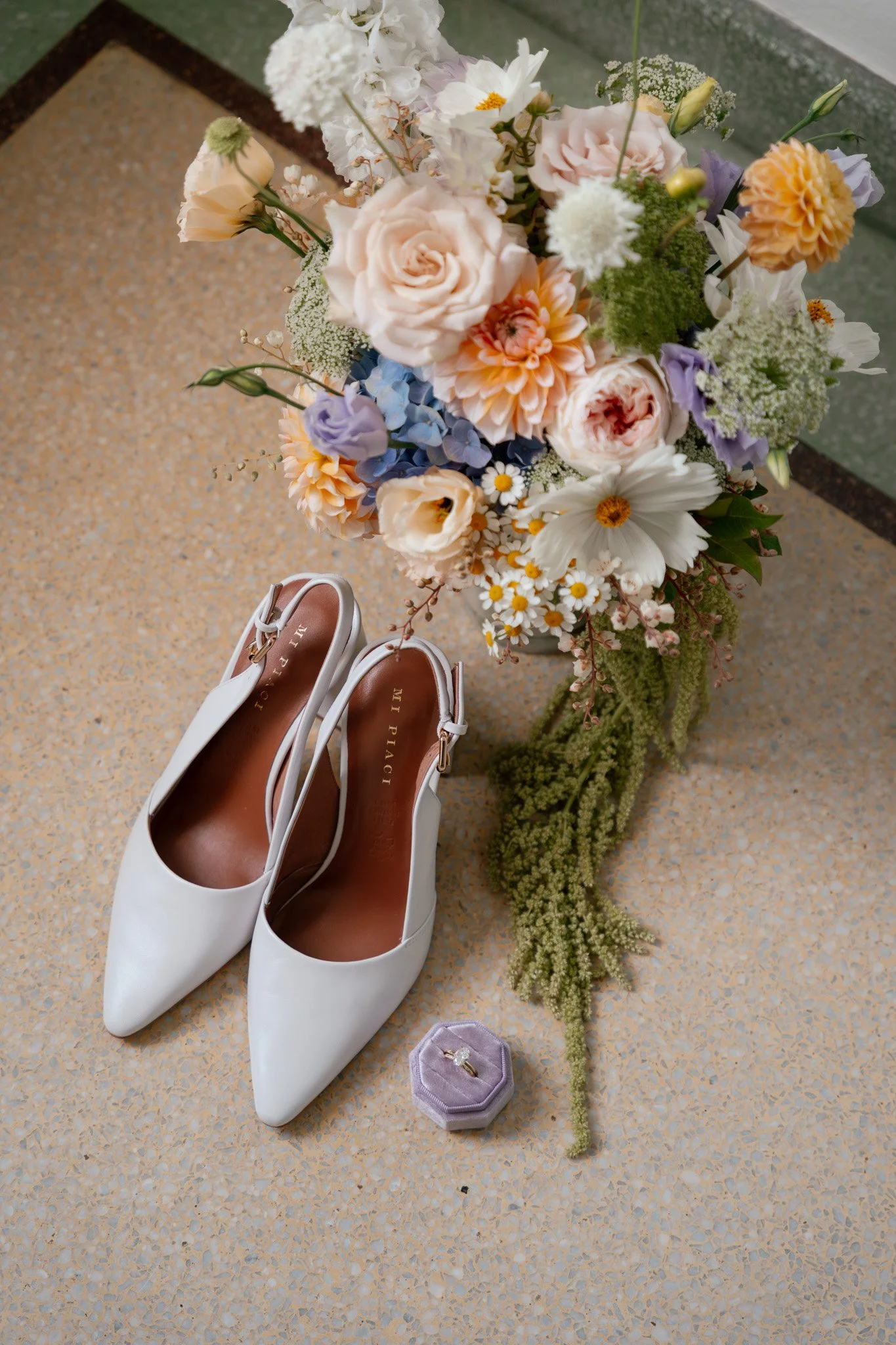 White high heel shoes, a bouquet of mixed pastel flowers including roses, daisies, and other blooms, and a lavender ring box with a diamond ring inside. All items are placed on a terrazzo floor.