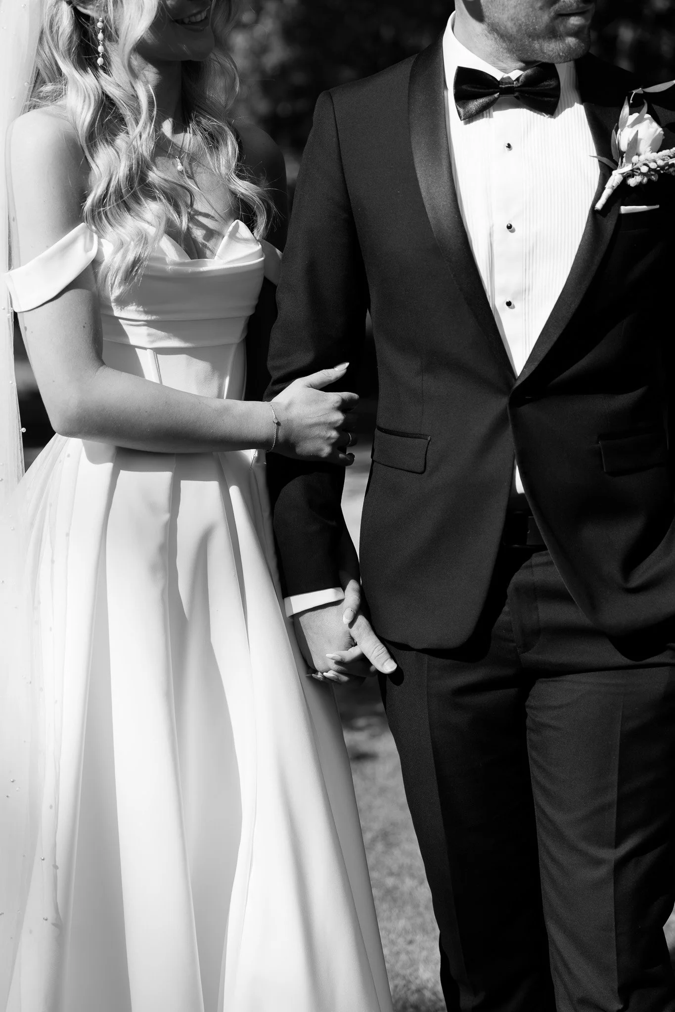 A bride and groom holding hands outdoors, the bride wearing an off-the-shoulder wedding gown and the groom in a tuxedo with a bow tie.