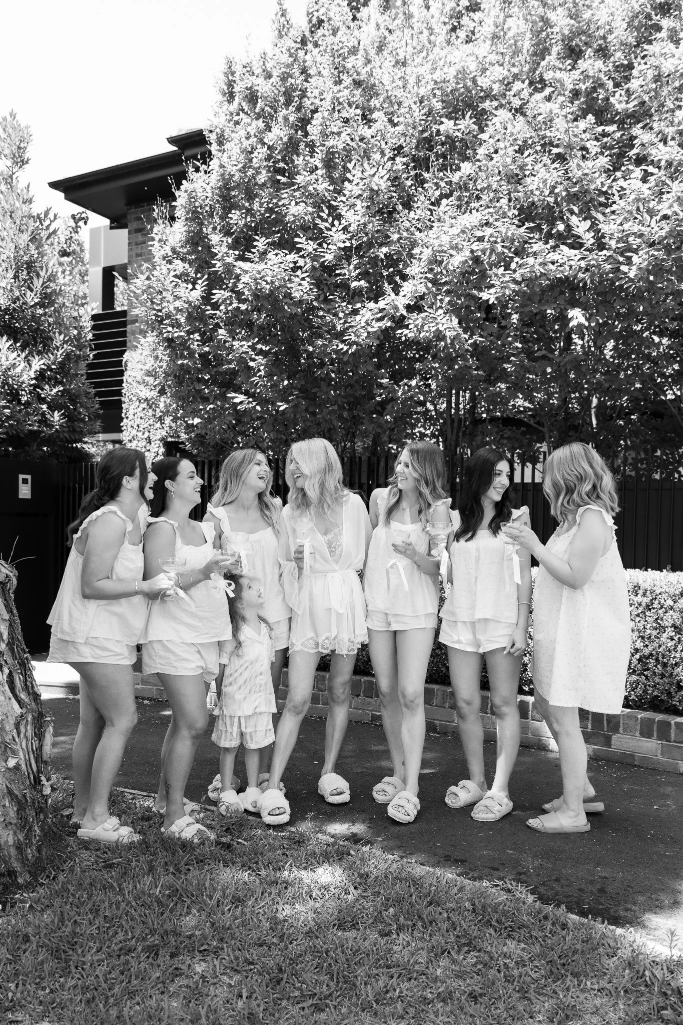 A group of women and a young girl in pajamas standing outdoors, smiling and holding drinks, in front of lush trees and a house.