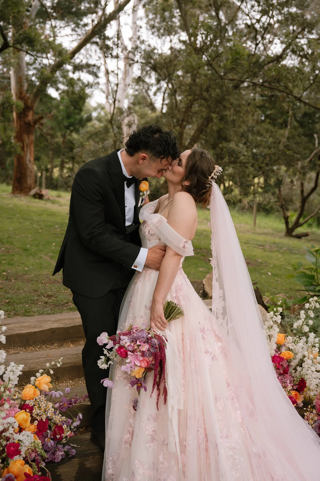 A newlywed couple kissing outdoors with trees and flowers around them. The bride is holding a bouquet of flowers and wearing a light pink wedding dress with a veil. The groom is dressed in a black tuxedo.