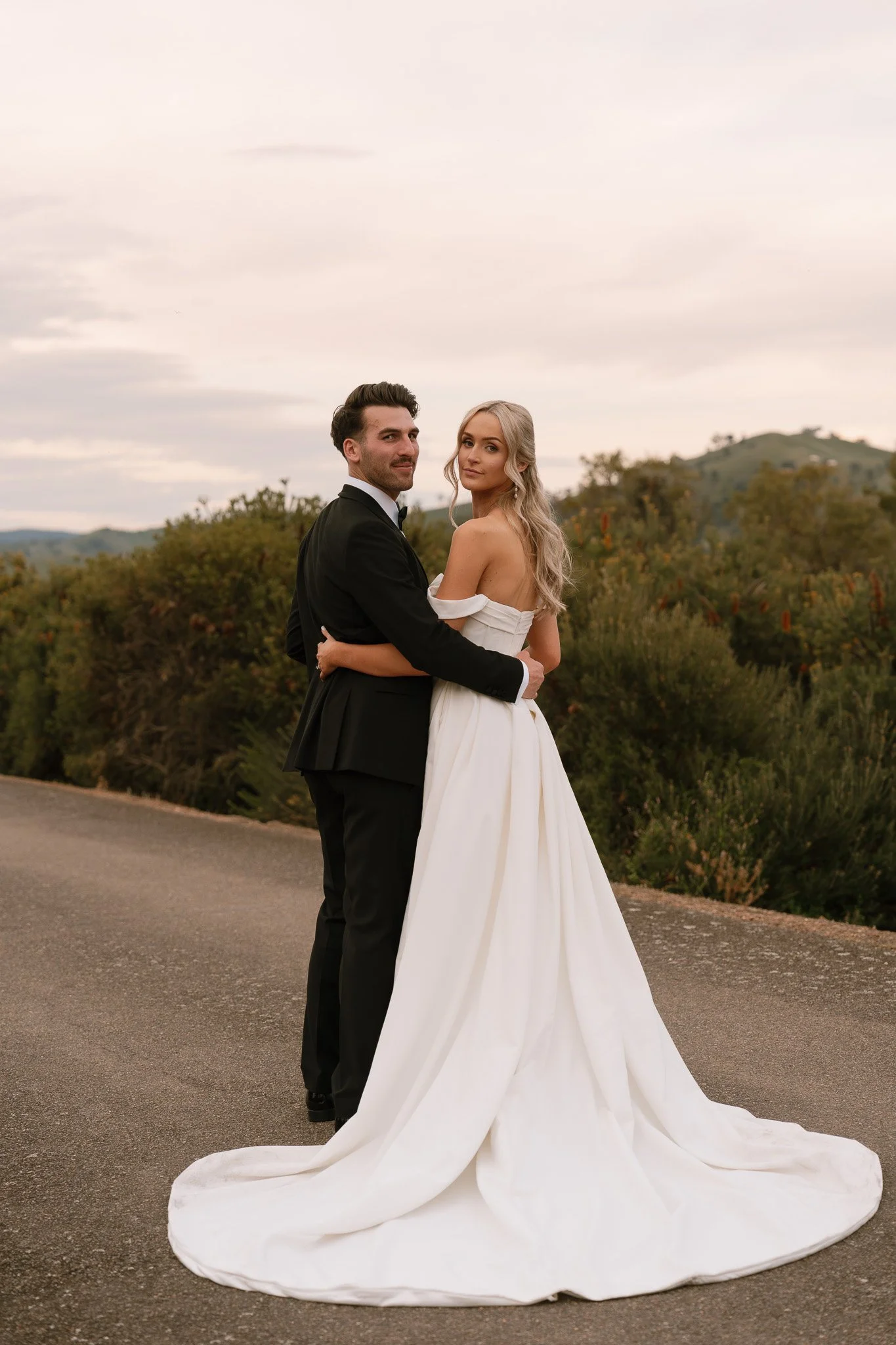A bride and groom standing together outdoors on a road, with the groom in a black tuxedo and the bride in a white off-the-shoulder wedding gown with a long train, surrounded by greenery and hills at sunset.