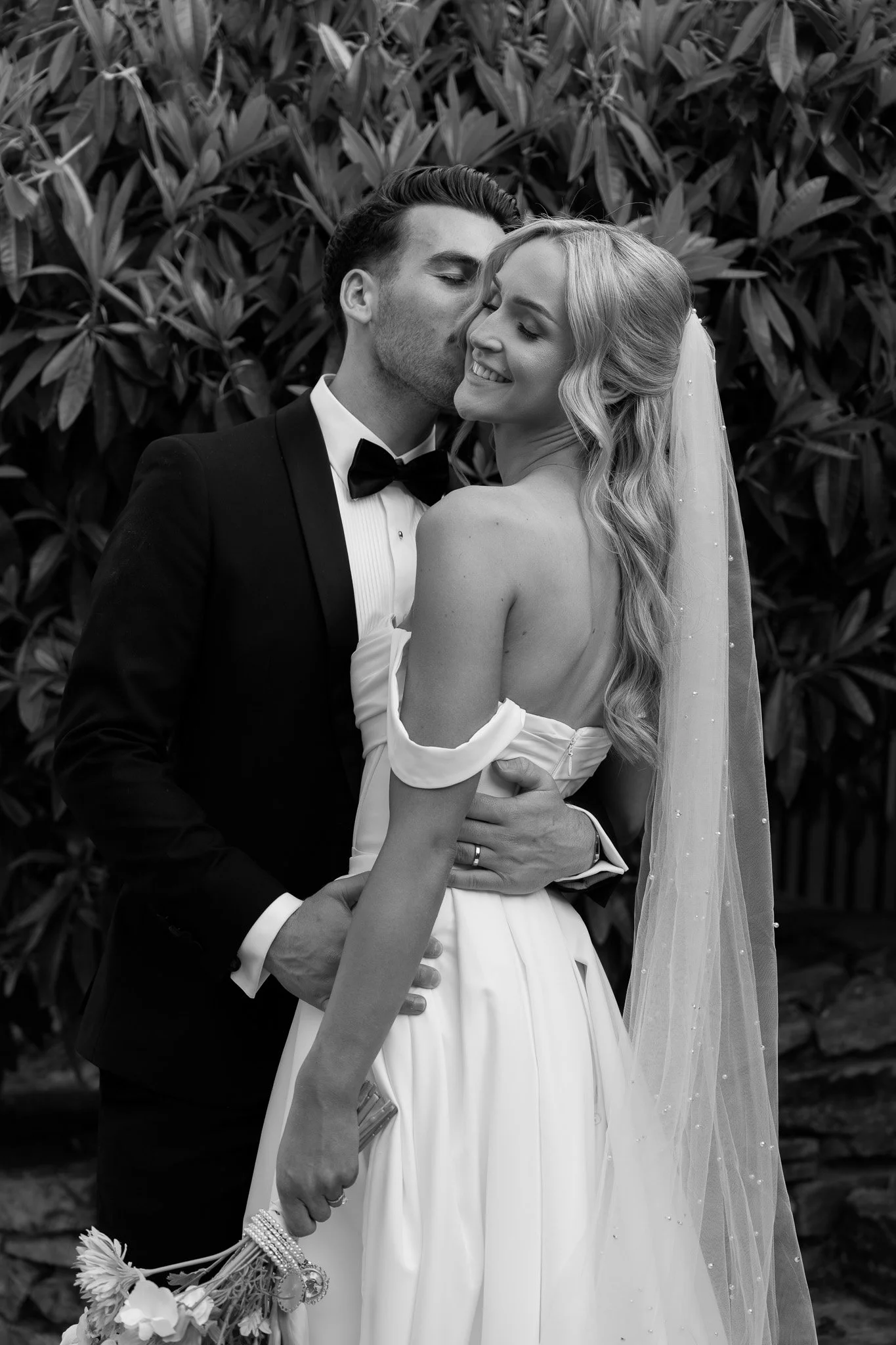 Black and white photo of a groom and bride on their wedding day, with the groom kissing the bride's temple and the bride smiling, holding a bouquet, with foliage in the background.