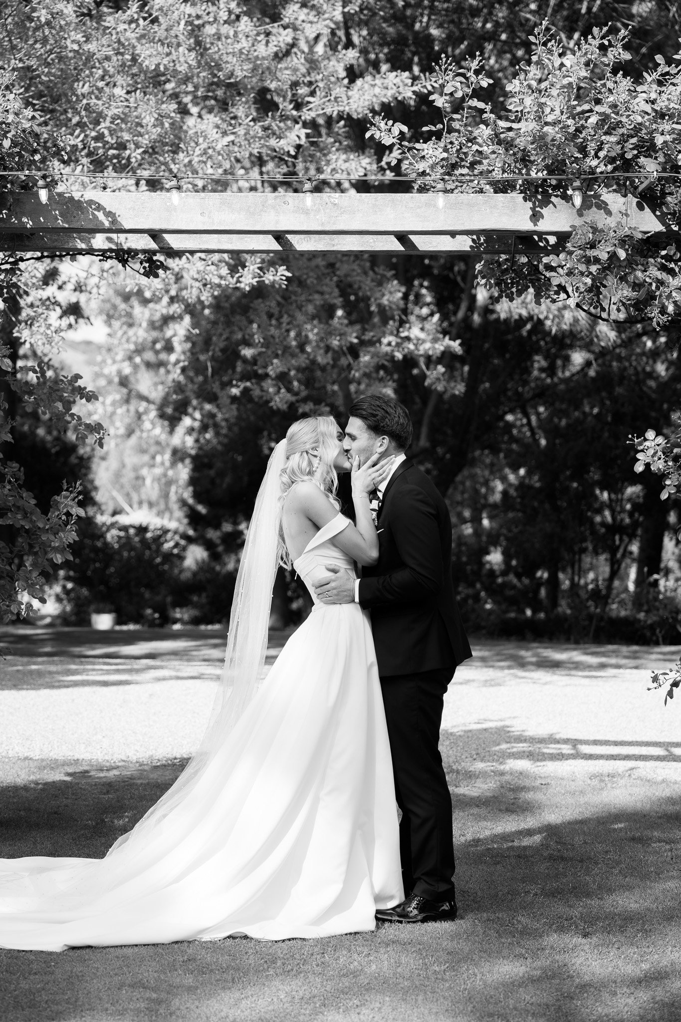 A bride and groom sharing a kiss outdoors, with trees and a string of lights overhead.