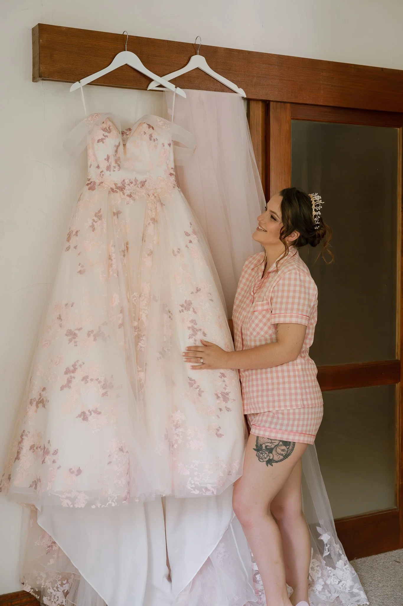 A woman in pink checkered pajamas looking at a wedding dress hanging on a wall with a sheer veil behind it.