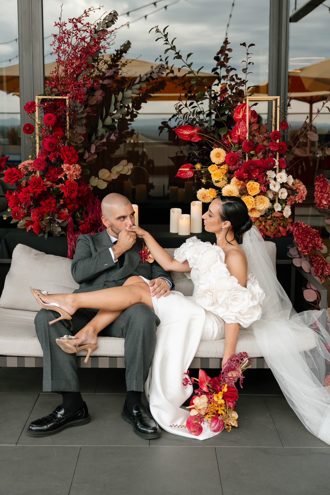 Bride and groom sitting on a sofa, with the groom kissing the bride's hand. The bride is holding a bouquet of red and yellow flowers, wearing a white dress with large floral details and a veil. The groom is in a gray suit with black shoes. Behind the