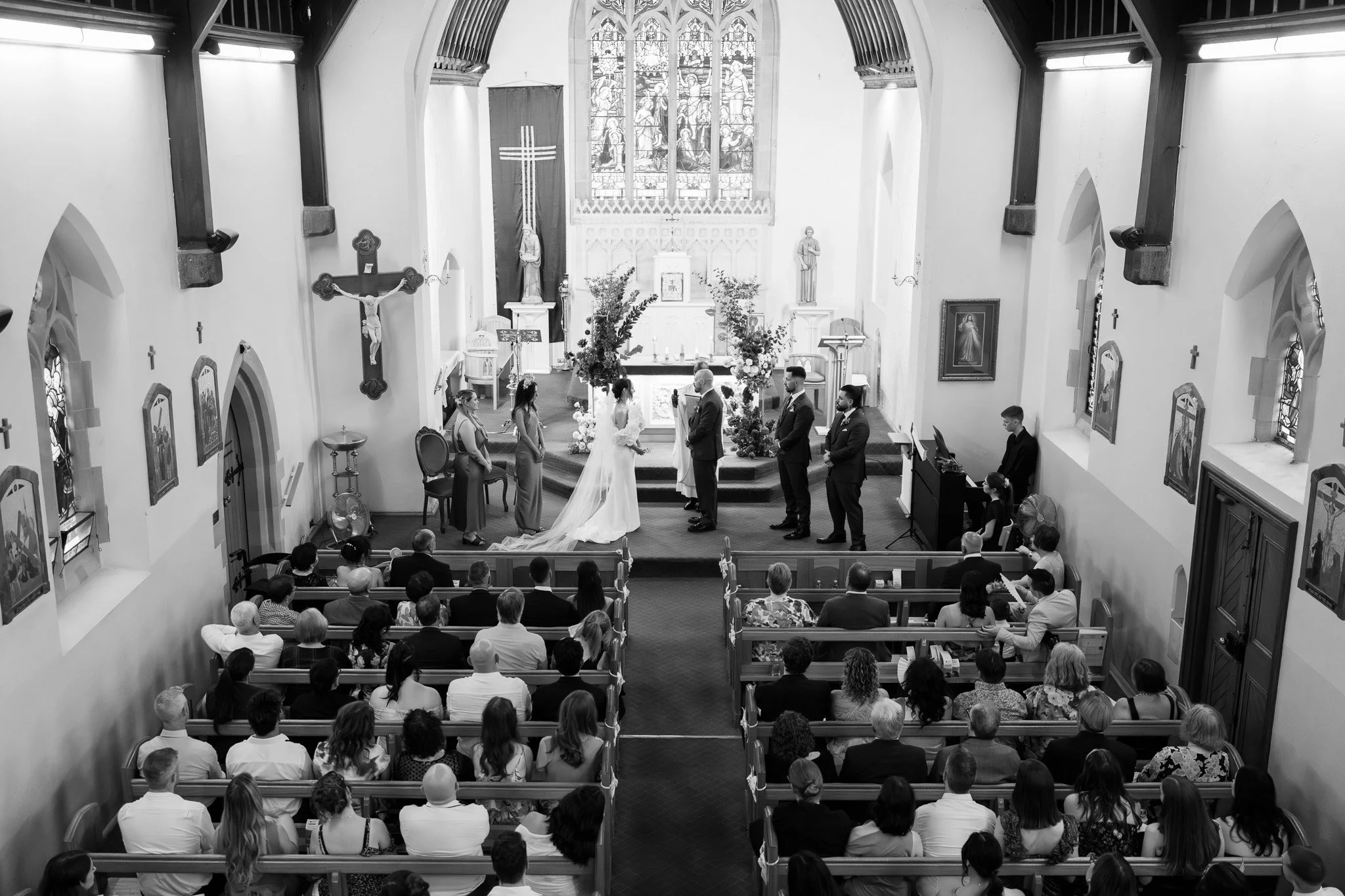 A wedding ceremony inside a church with the bride and groom standing at the altar, surrounded by family, friends, and clergy, with stained glass windows and religious statues in the background.