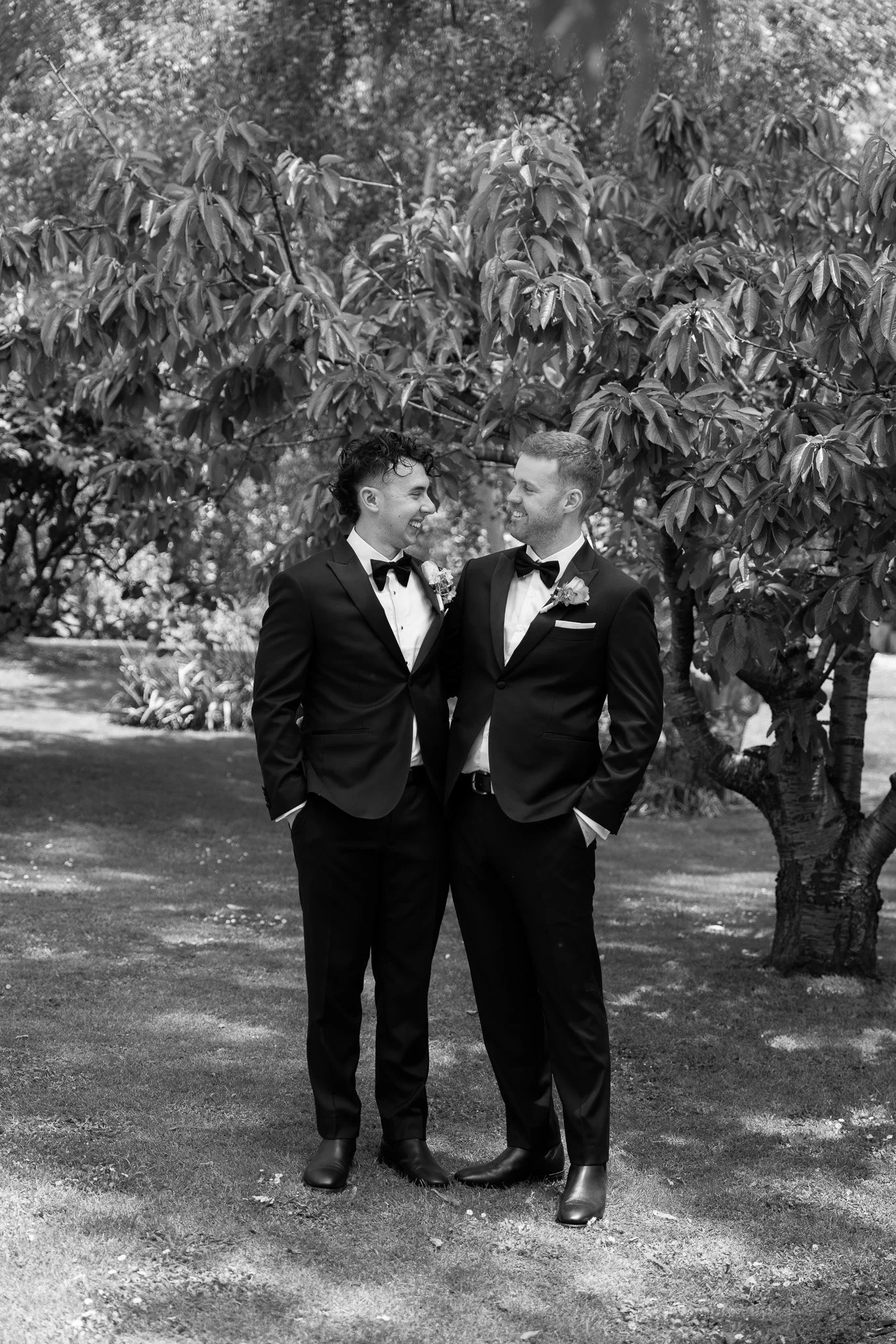 Black and white photograph of two men in tuxedos standing close together outdoors, smiling at each other, with a large leafy tree in the background.