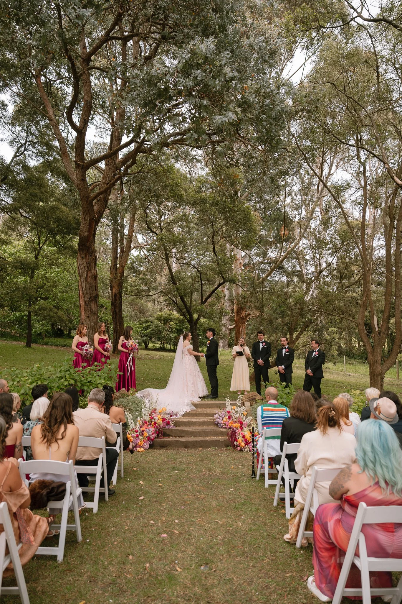 Outdoor wedding ceremony with bride and groom exchanging vows, officiant, five groomsmen, five bridesmaids, seated guests, and floral decorations in a wooded area.