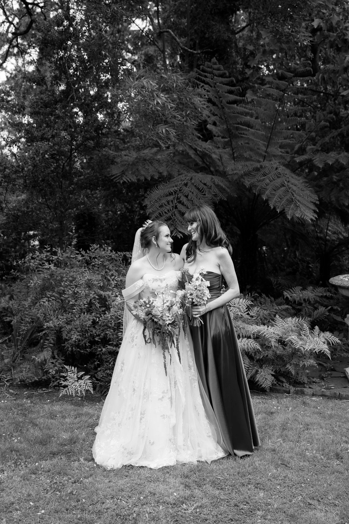 Two women, one in a wedding dress and the other in a dark strapless gown, standing on grass in front of large leafy plants, holding bouquets, smiling at each other.