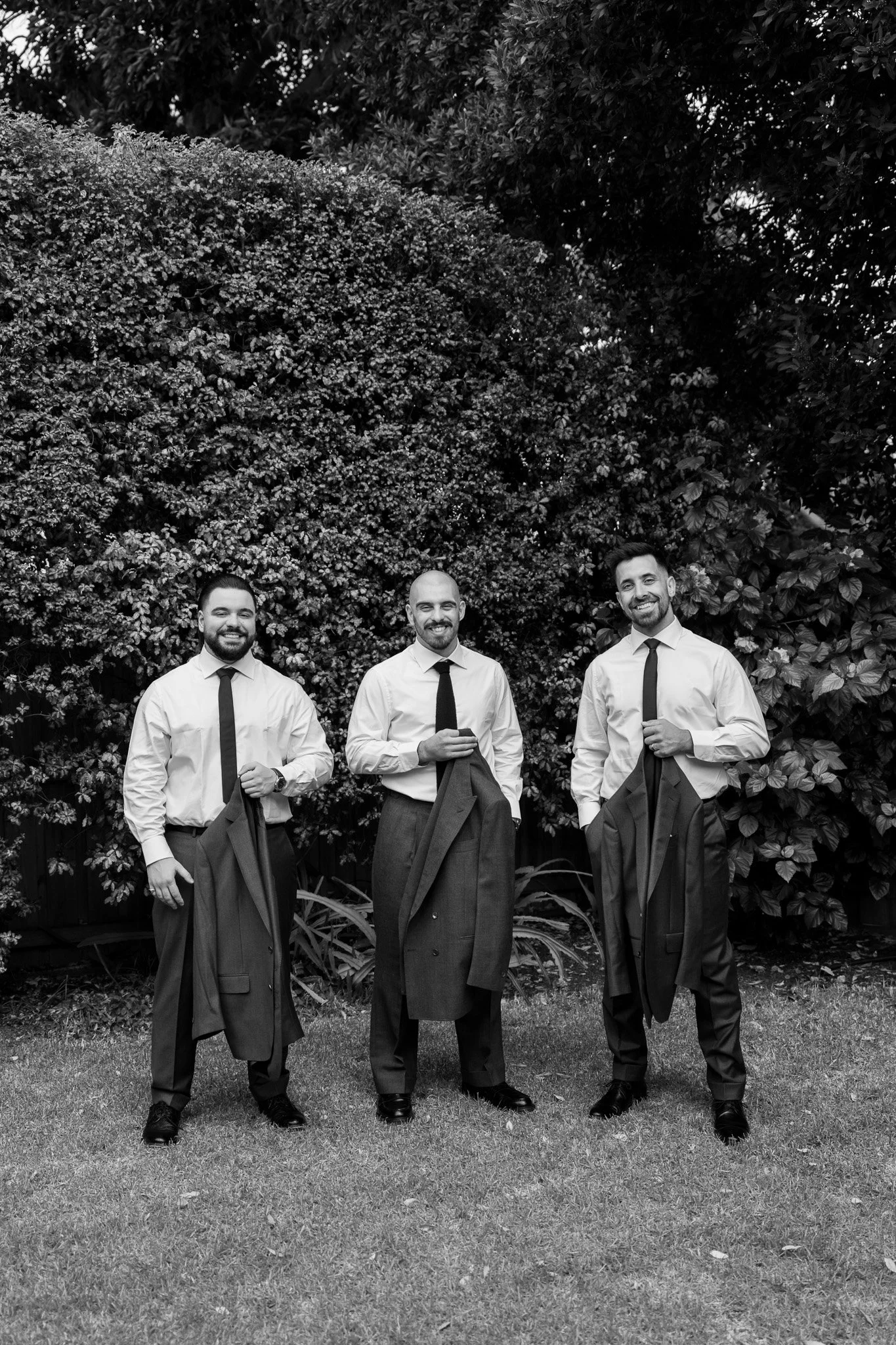 Three men dressed in white shirts, black ties, and black trousers, holding their jackets, standing on a lawn in front of shrubbery with trees in the background, smiling.