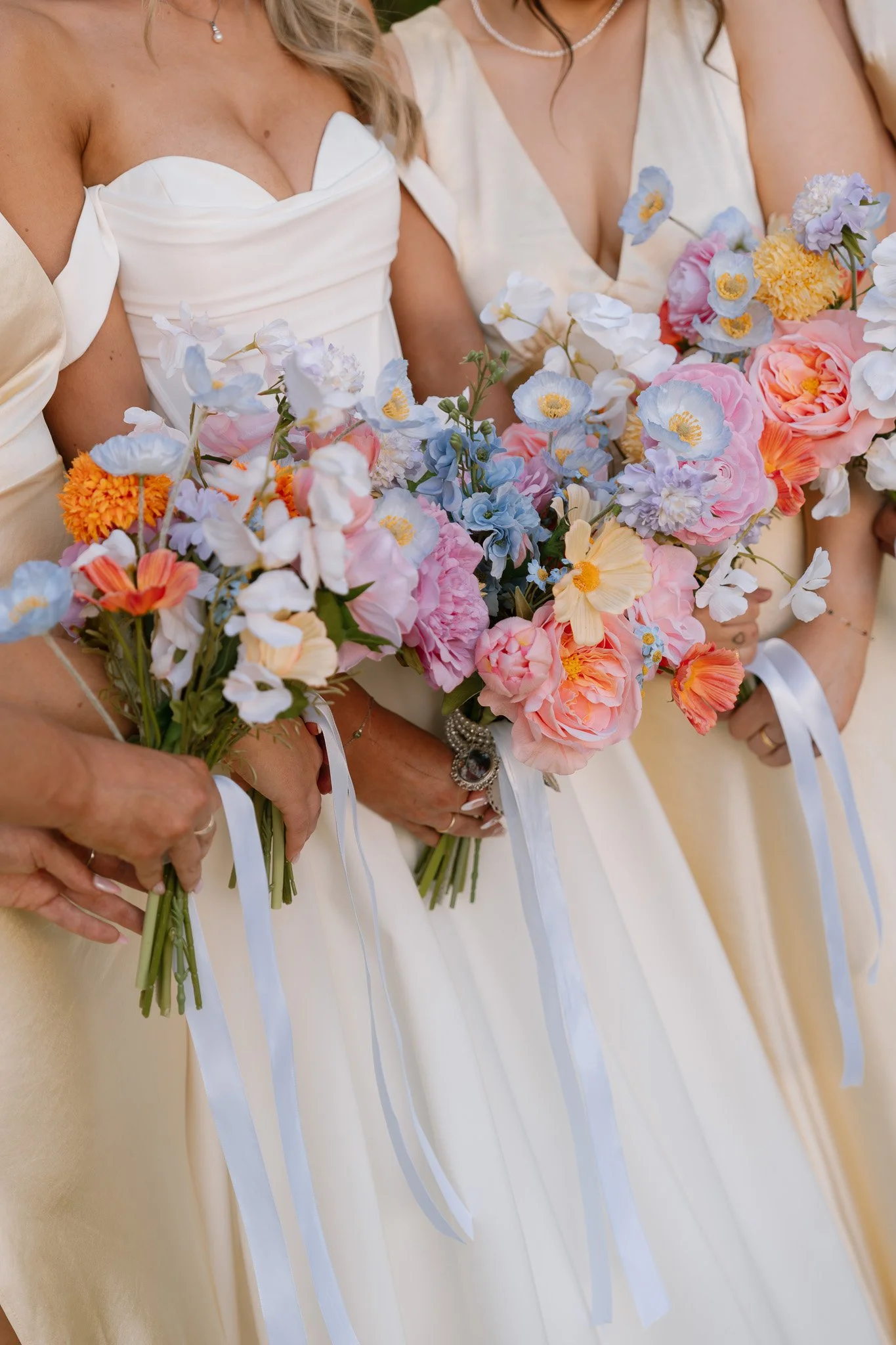 Women in cream-colored dresses holding bouquets of pink, purple, white, and orange flowers, with white ribbon ties, at a wedding or formal event.