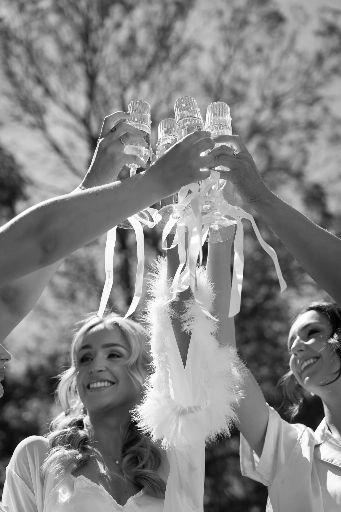 Three women raising glasses in a celebratory toast outdoors, with trees in the background, in black and white.
