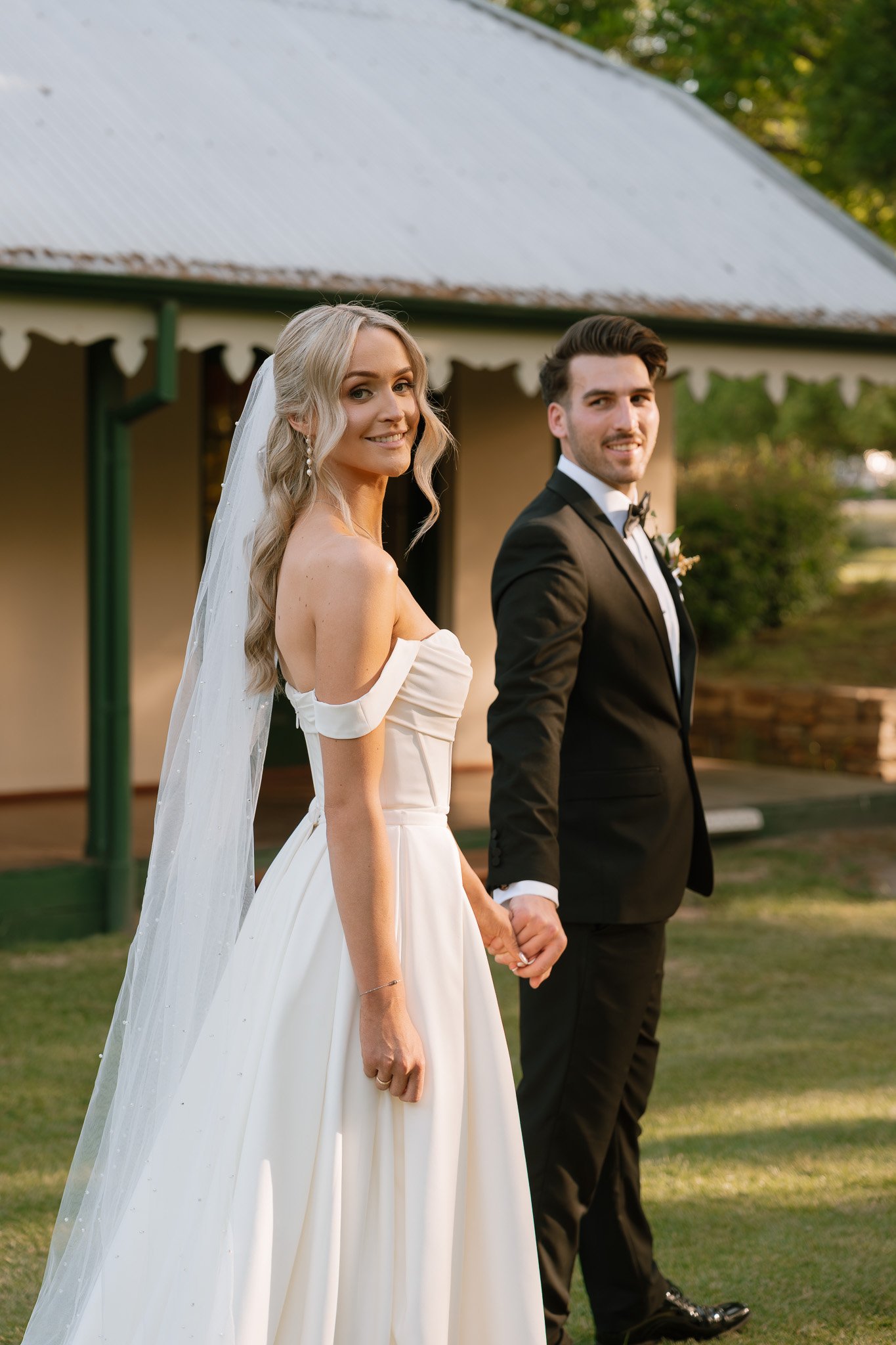 A bride and groom holding hands outdoors during their wedding, standing on a grassy area in front of a building with a peaked roof.
