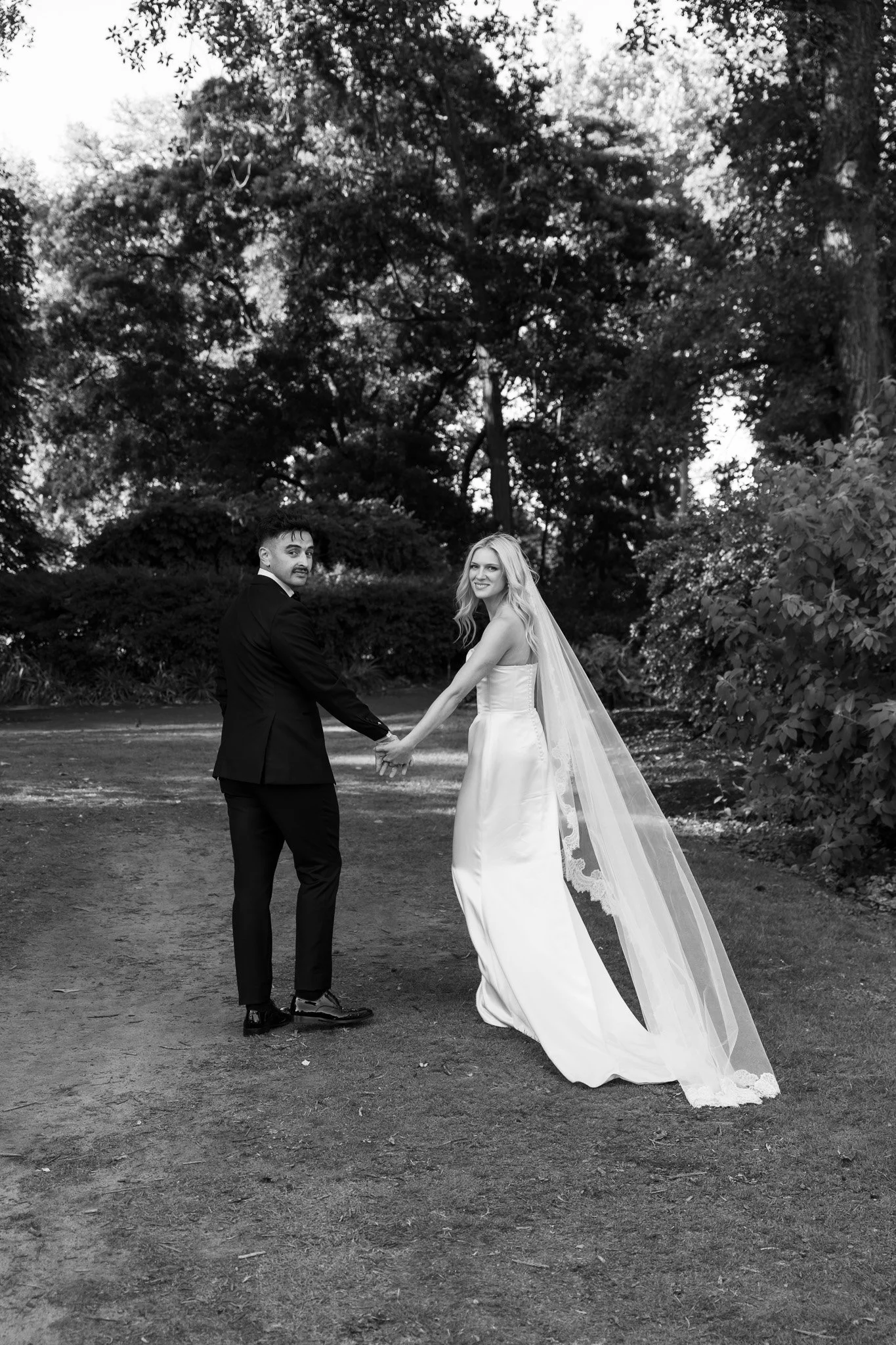 Black and white photo of a newlywed couple holding hands outdoors, with the groom in a suit and the bride in a wedding dress and veil, standing on a dirt path surrounded by trees and bushes.