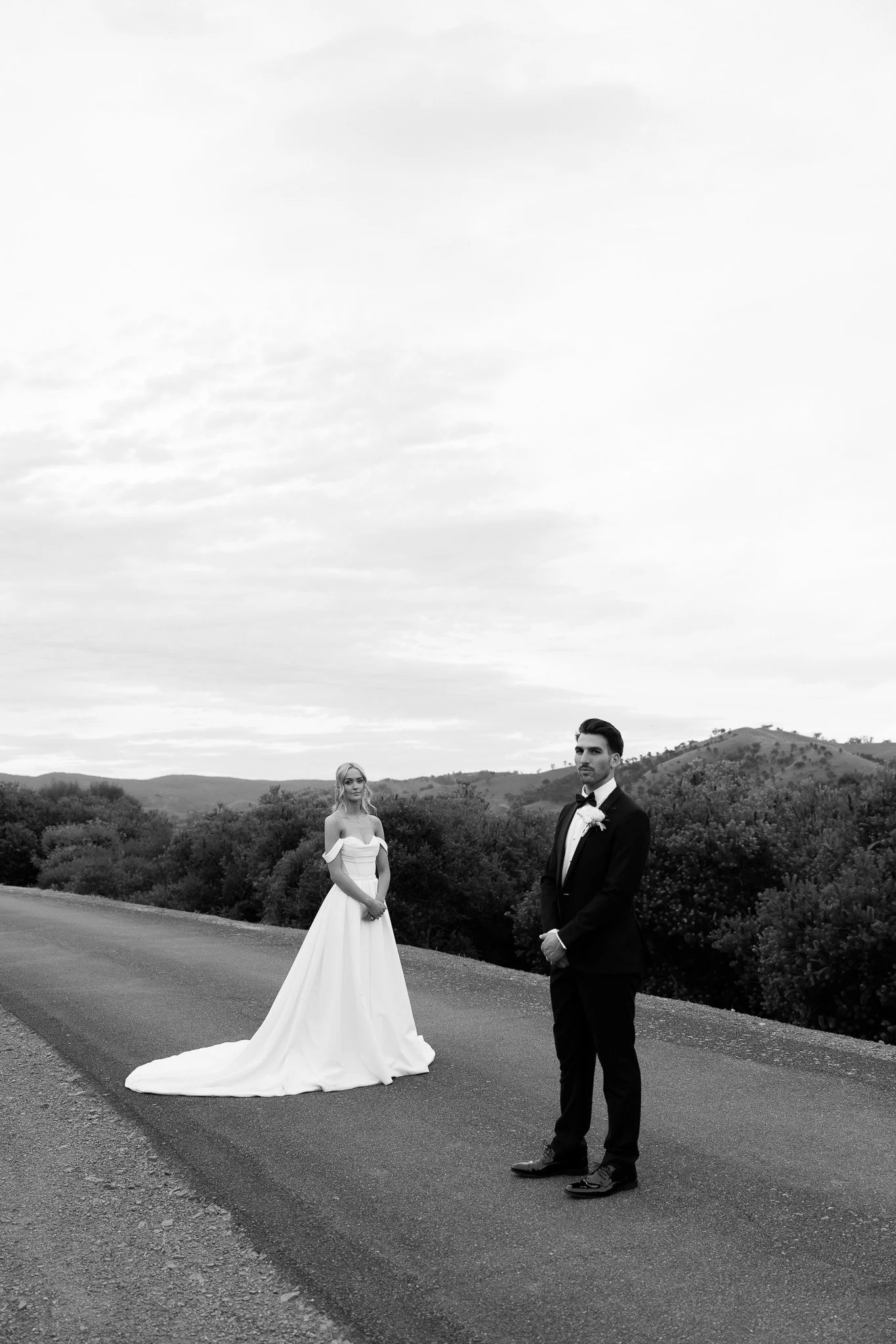A black and white photo of a bride and groom standing on a rural road with hills and bushes in the background. The bride is wearing a white wedding gown with off-shoulder sleeves, and the groom is in a black tuxedo with a bow tie.