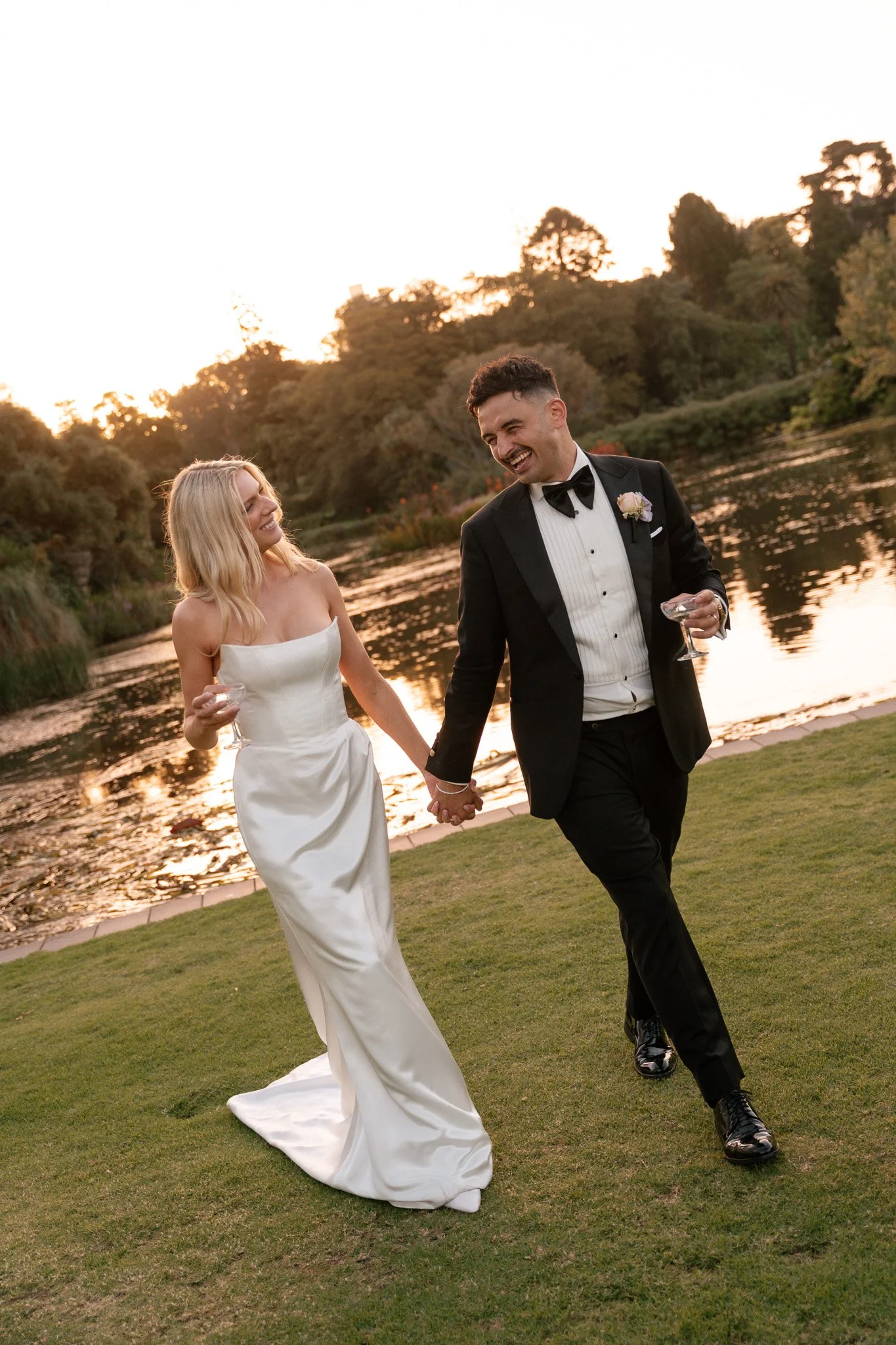 A newlywed couple walking hand-in-hand by a lake during sunset, both holding champagne glasses and smiling happily.