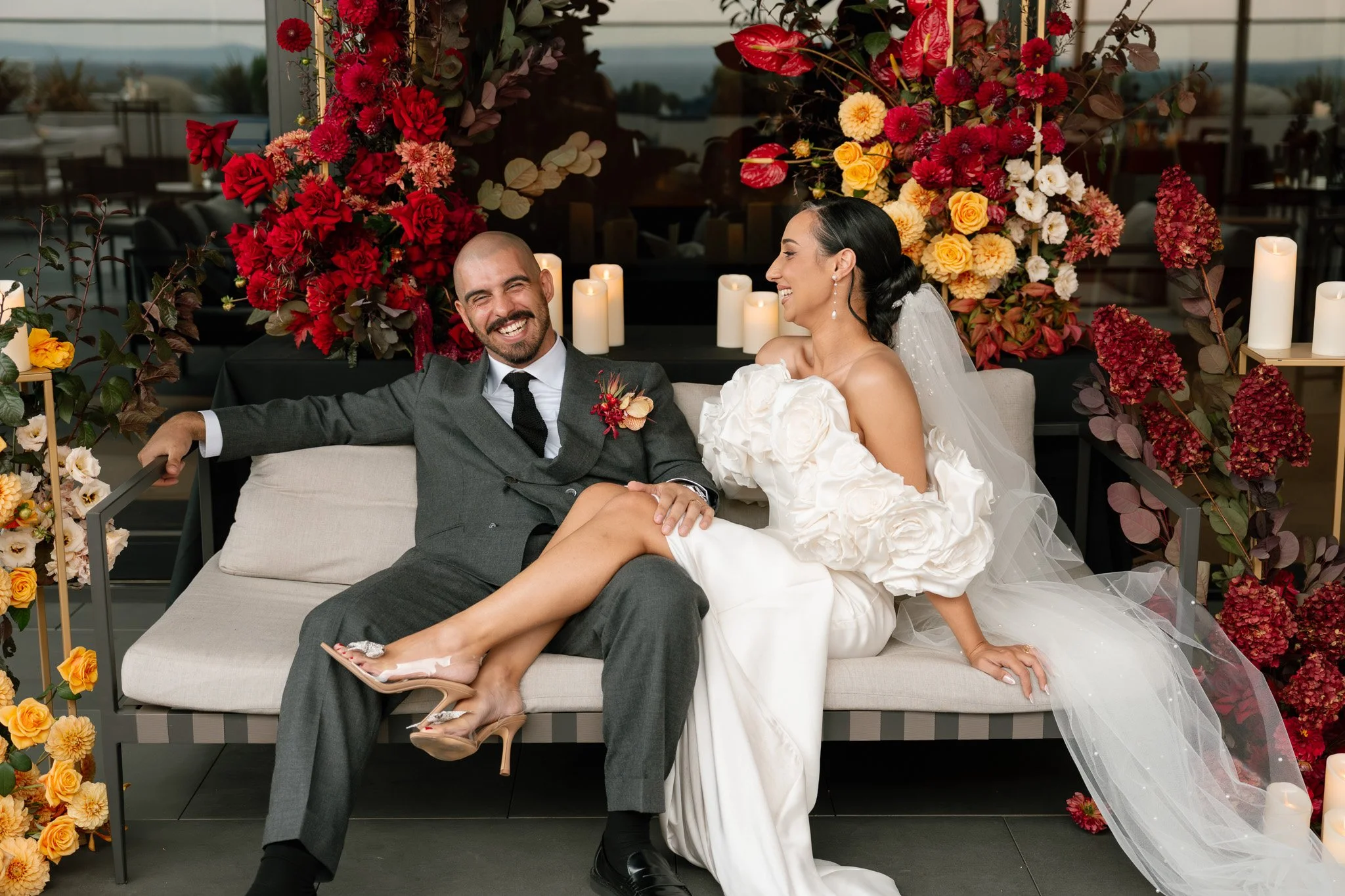 A happy bride and groom sitting on a light-colored outdoor sofa surrounded by tall red, pink, and yellow flowers and lit candles, sharing a joyful moment.