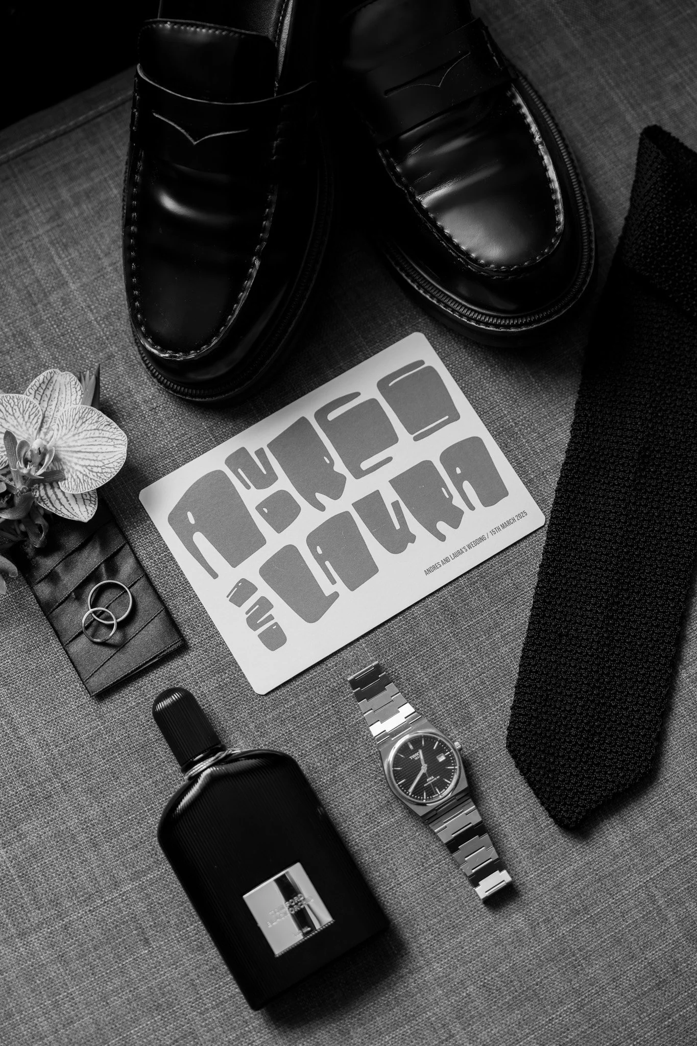 Black leather shoes, a wristwatch with a metal band, a bottle of cologne, a pair of wedding rings, a small potted orchid, a black textured tie, and a wedding invitation card on a dark fabric surface.