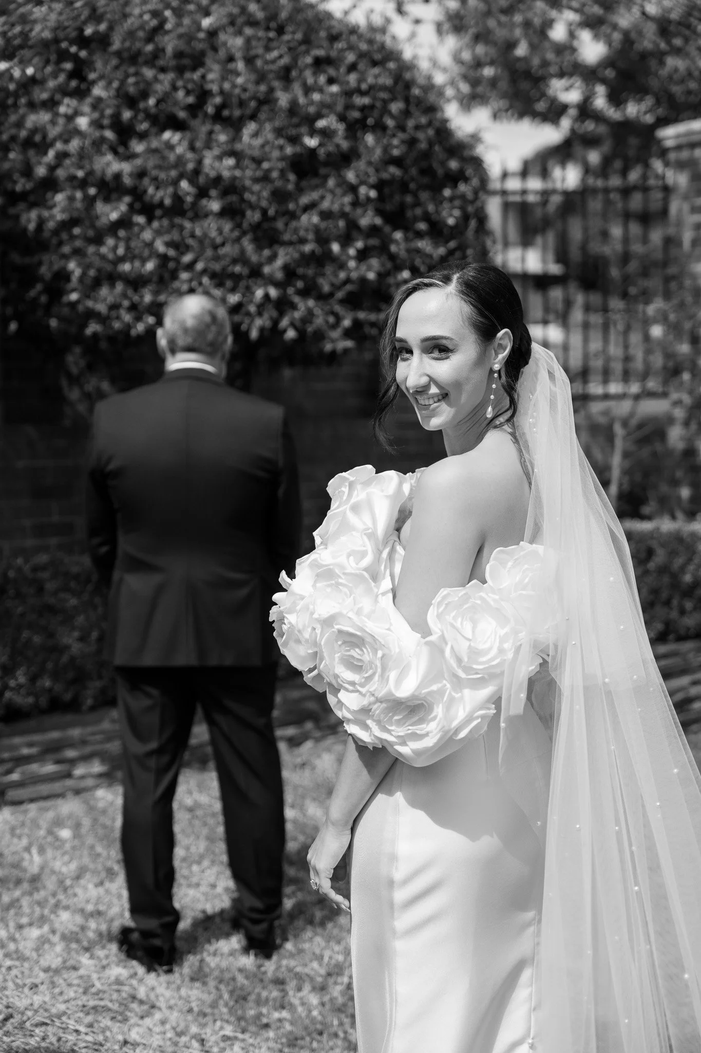 A bride in a wedding dress and veil smiling at the camera, standing outdoors, with an older man in a suit facing away in the background.
