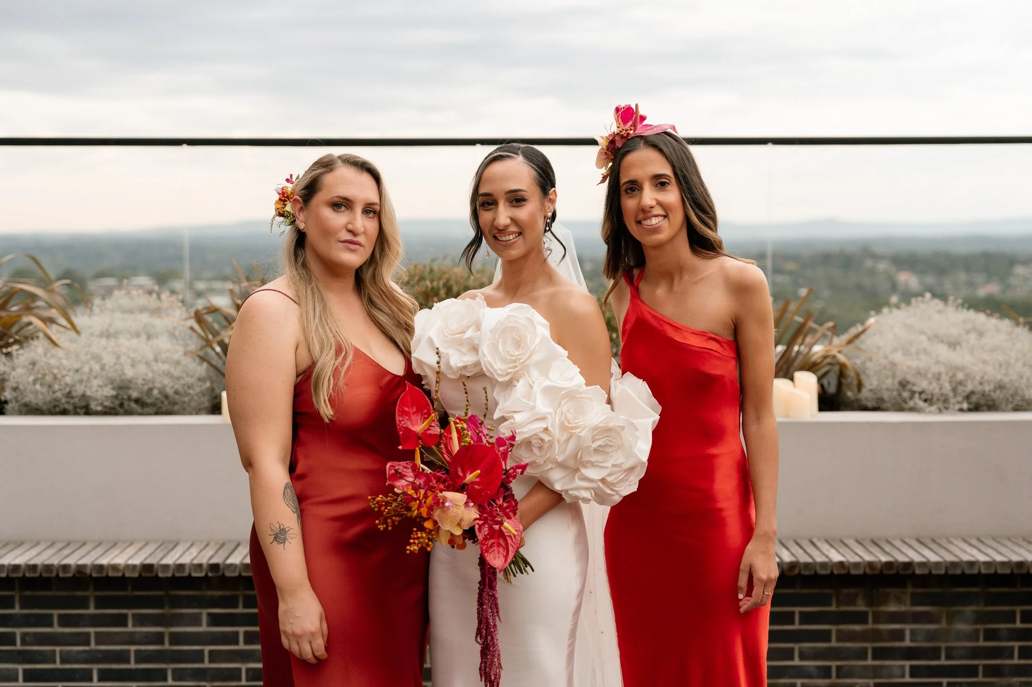 Three women on a rooftop, with the bride in the middle wearing a white wedding dress holding a bouquet of large white and red flowers, and two bridesmaids in red dresses on either side, with plants and a city view in the background.