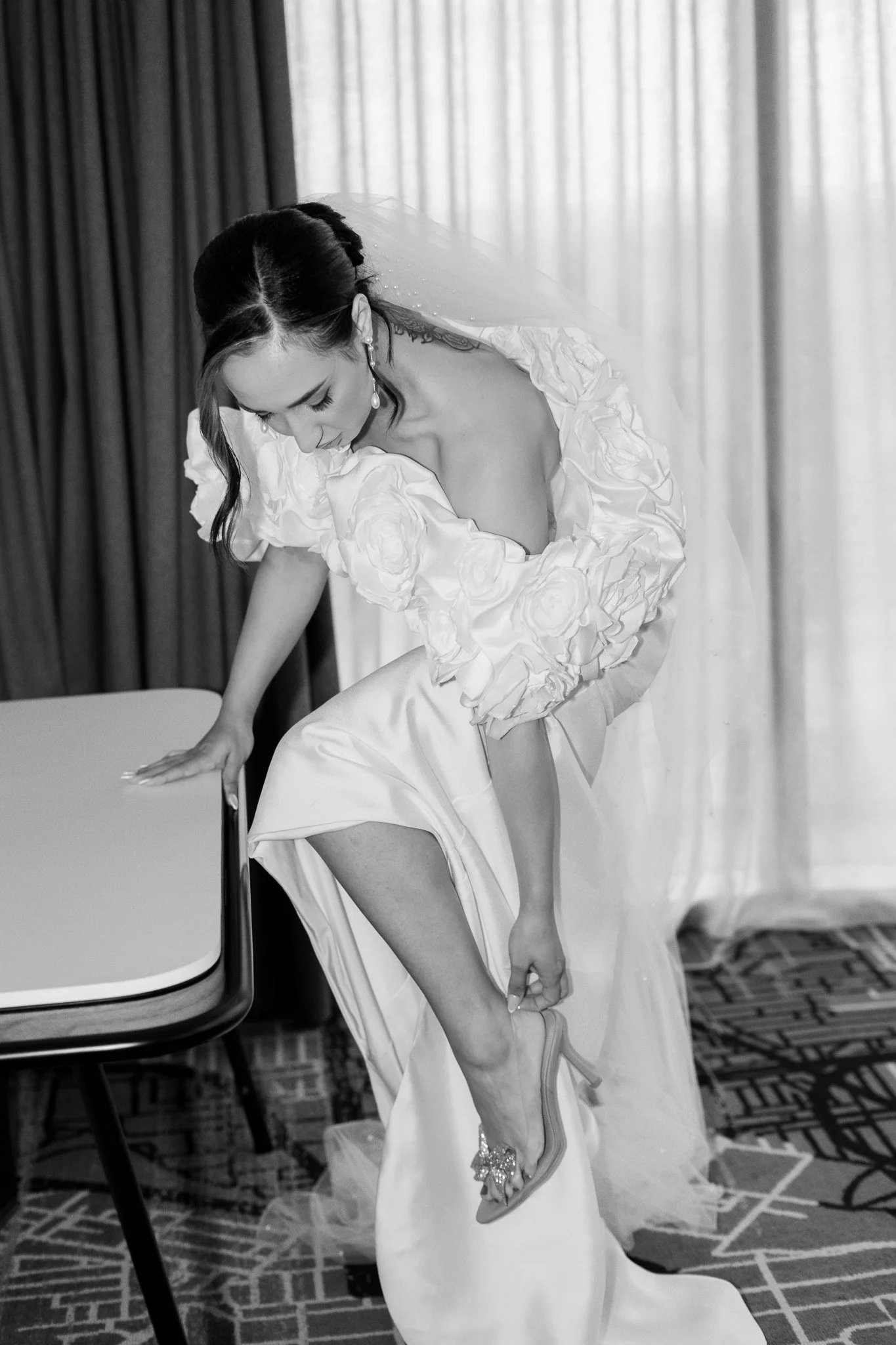 Black and white photo of a bride in a wedding dress leaning down, adjusting her high heel shoe, with a floral design on the dress's sleeves and a veil behind her.