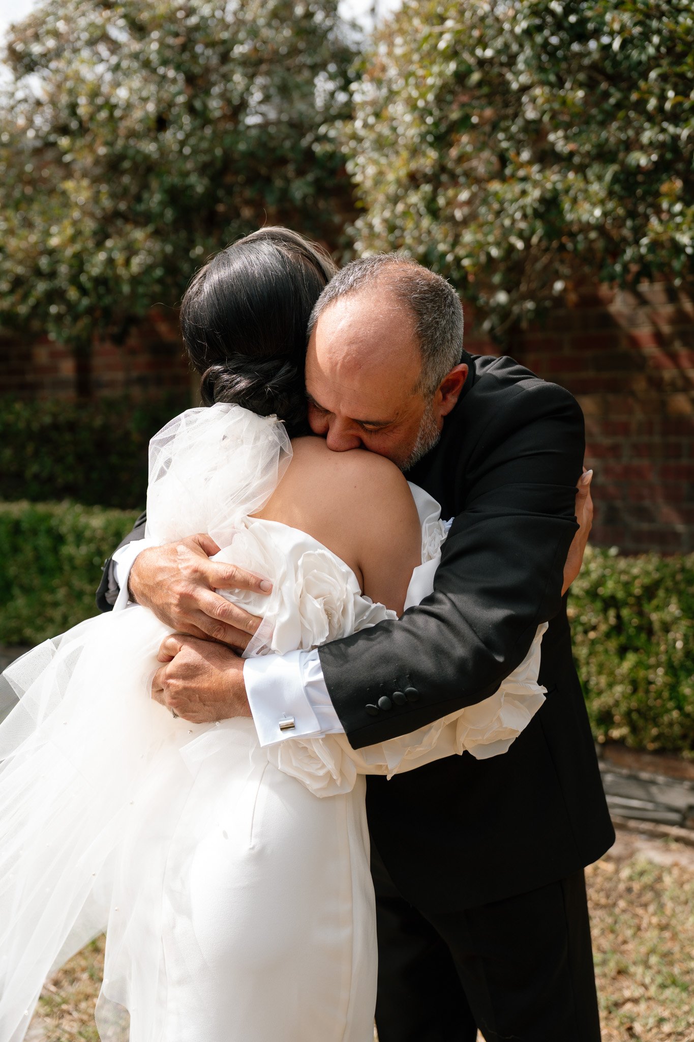 A man in a black suit hugging and kissing a woman in a wedding dress outdoors.