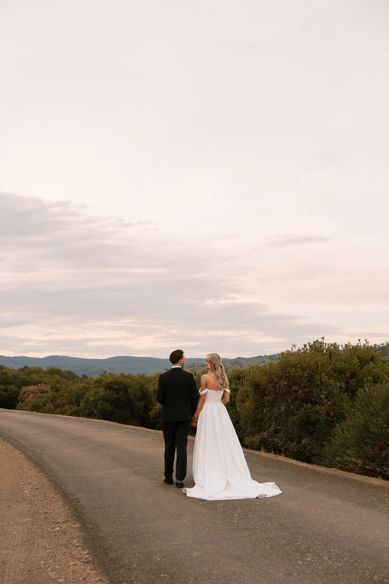 A bride and groom walking hand in hand on a country road with hills and trees in the background.