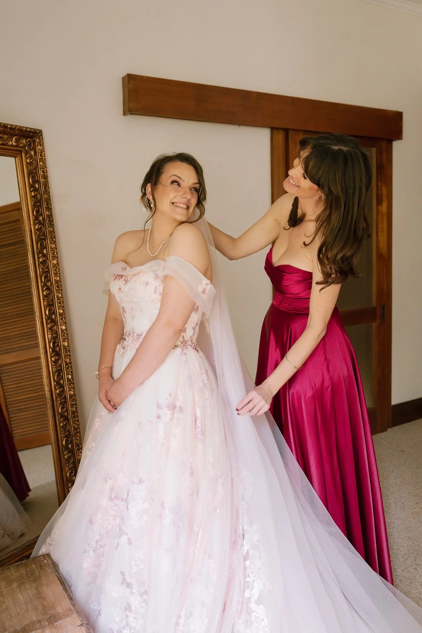 A bride with a floral off-shoulder wedding gown smiling as her bridesmaid in a red satin dress helps her with the dress.