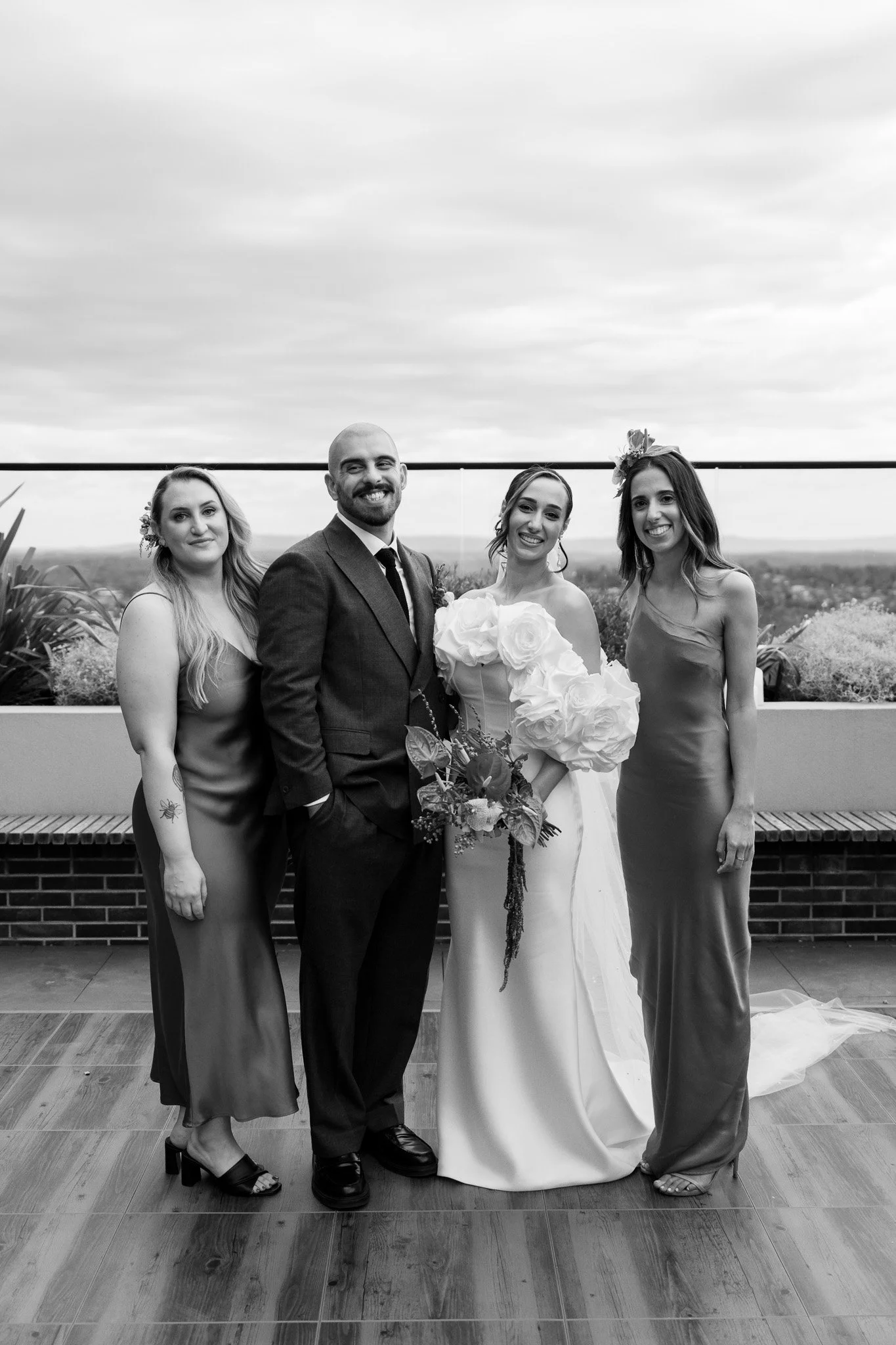 Black and white photo of four people at a wedding, with two women, a man, and a bride holding a bouquet, standing on a wooden floor with a scenic outdoor view in the background.