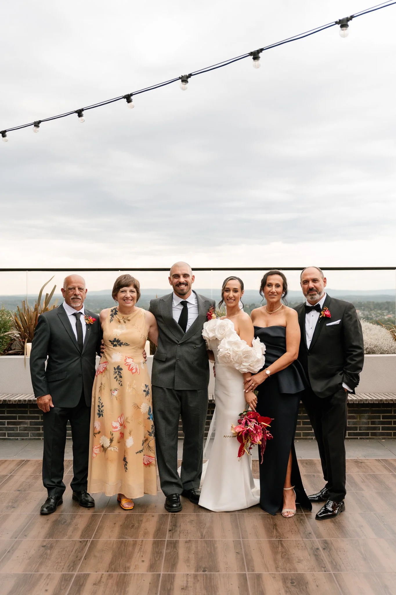 Group of six people in formal attire on a rooftop, with wedding couple in the center, overcast sky in the background.