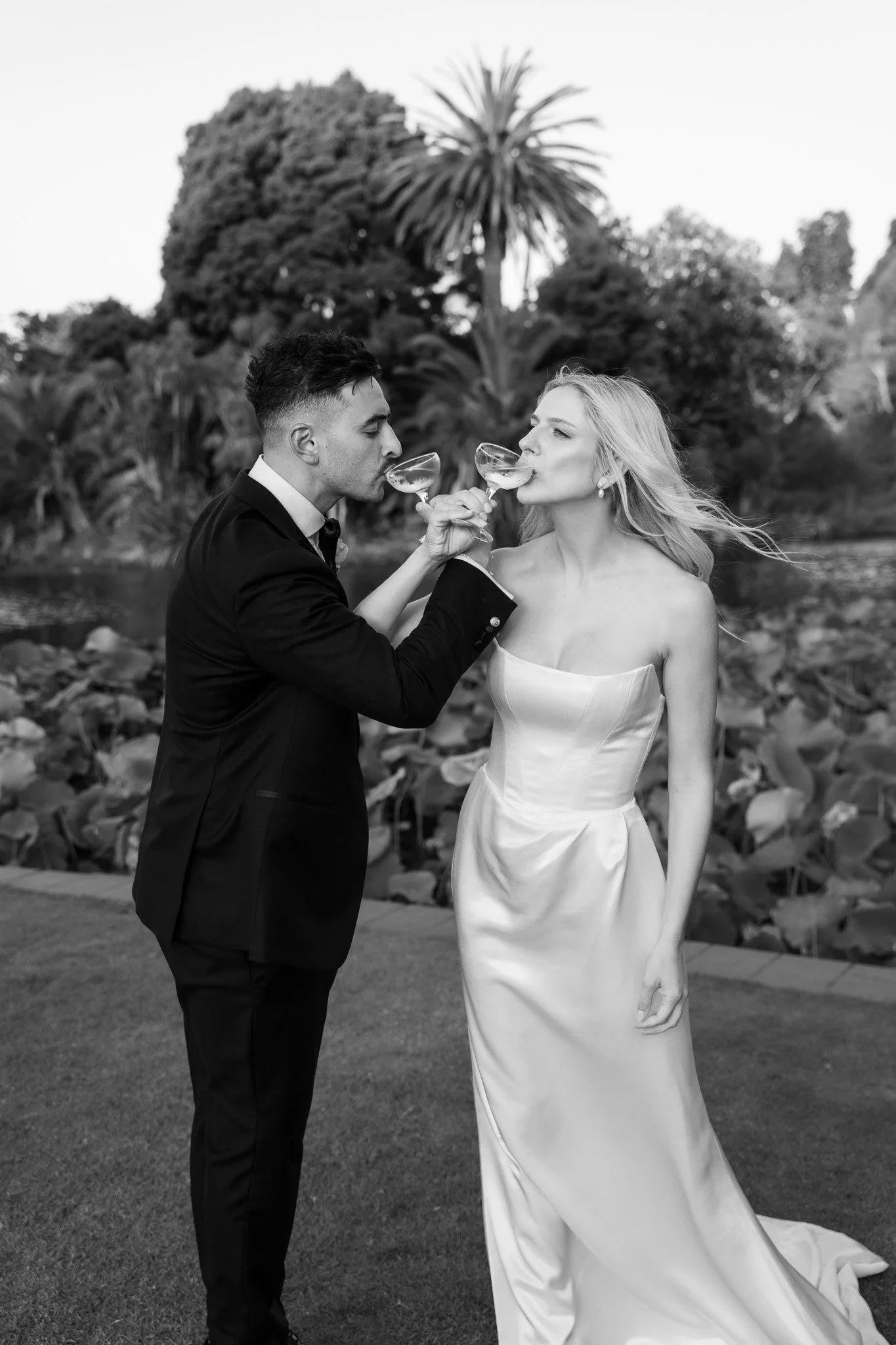 A black and white photo of a couple in formal attire sharing a toast with champagne glasses outdoors near water, with trees and large plants in the background.
