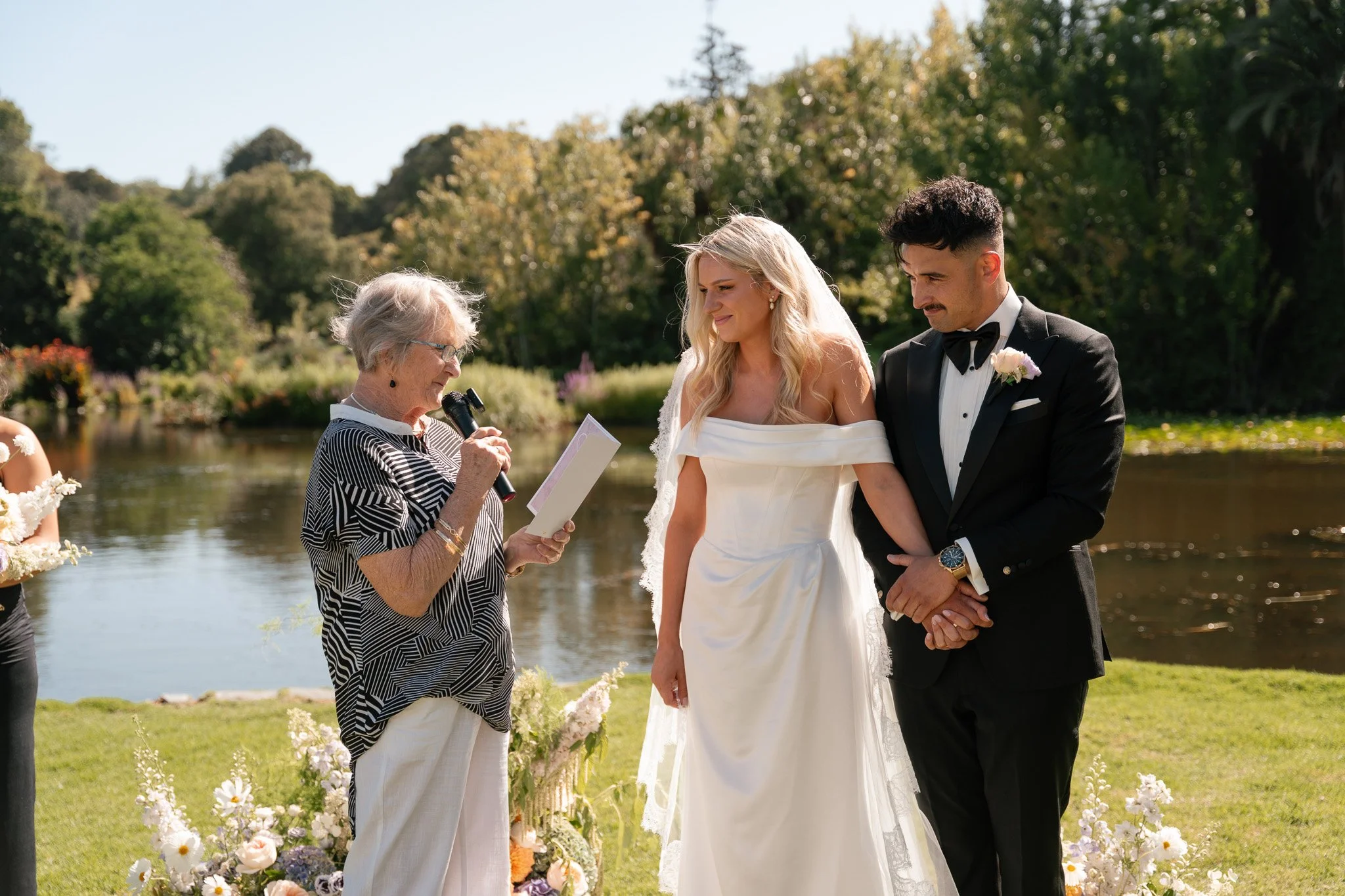 A couple getting married outdoors, standing beside an officiant who is reading from a paper, with a river and green trees in the background. The bride is in a white off-shoulder wedding dress, and the groom is in a black tuxedo with a bow tie. They a