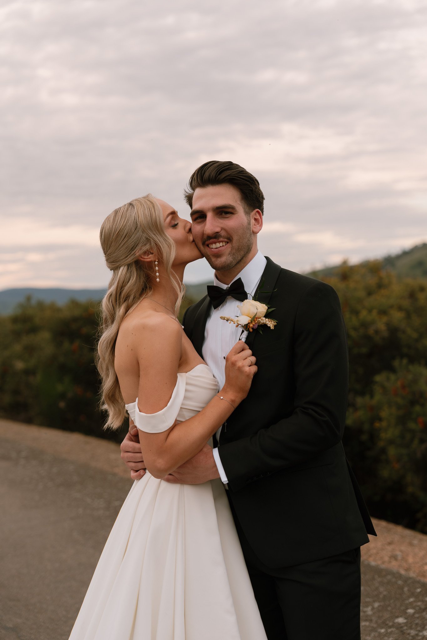 A bride and groom embracing outdoors on their wedding day, with the bride kissing the groom's cheek as he smiles, wearing a black tuxedo, against a cloudy sky and scenic landscape.