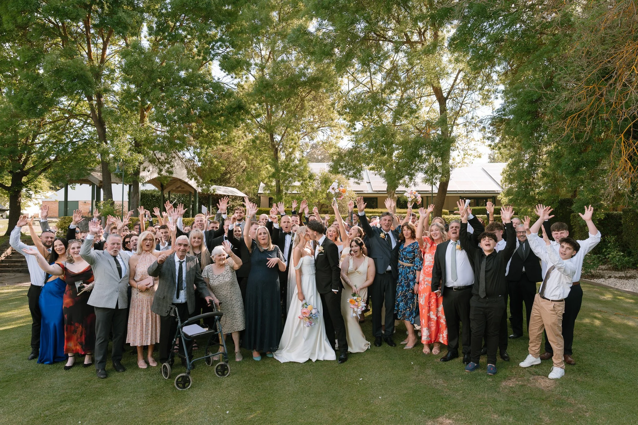 Group of wedding guests outdoors on a grassy lawn, celebrating with raised hands, in front of trees and a building.