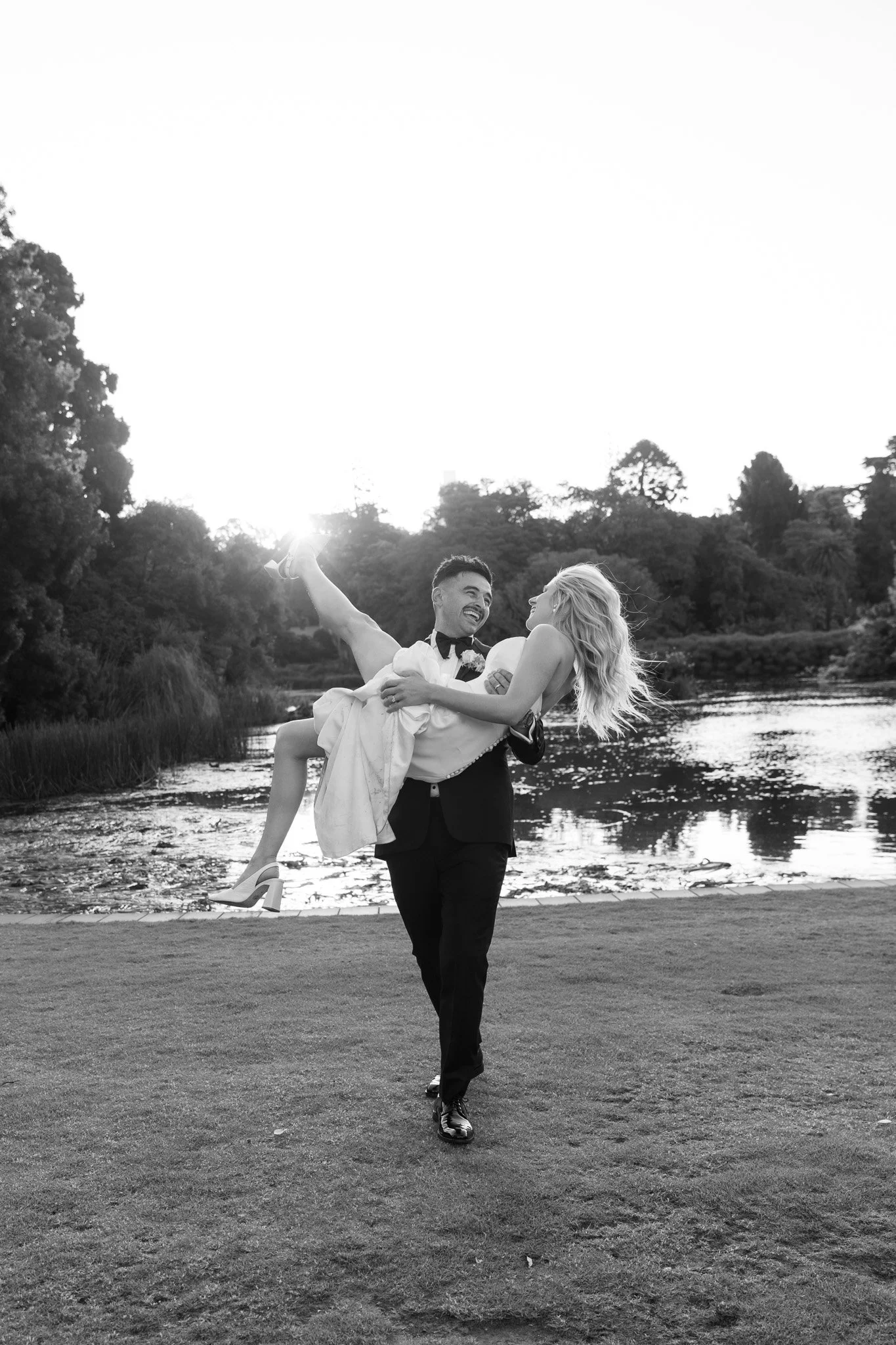 A groom in a tuxedo is holding a bride in a wedding dress and high heels, lifting her as they dance outdoors by a lake, with trees and the setting sun in the background.