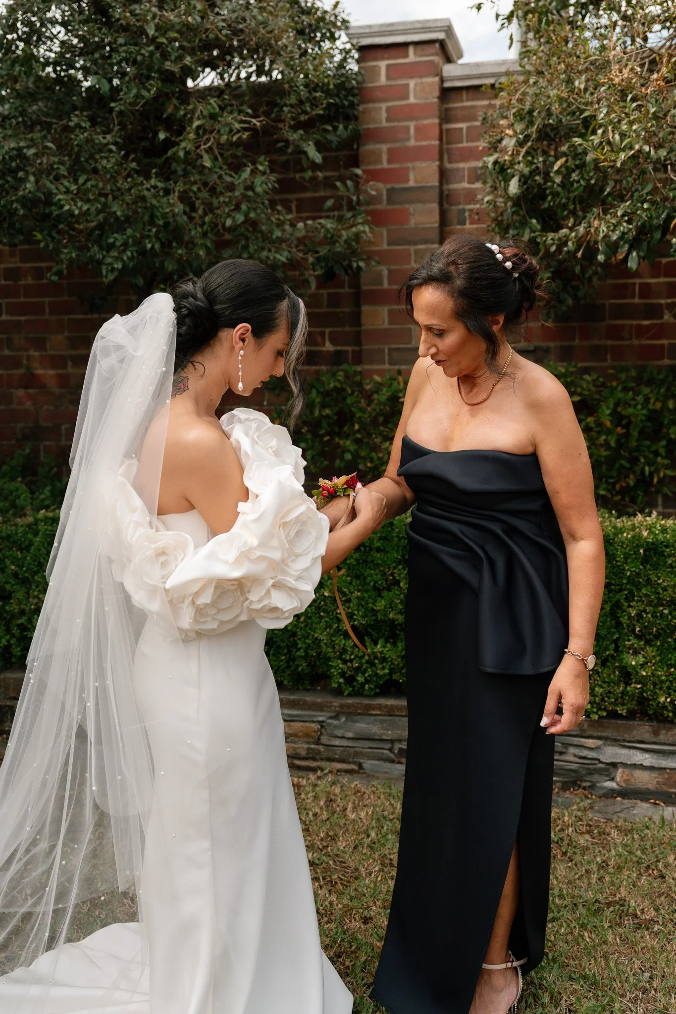 A bride in a wedding dress and veil is holding hands with a woman in a black dress during a wedding ceremony outdoors, with a brick wall and green bushes in the background.