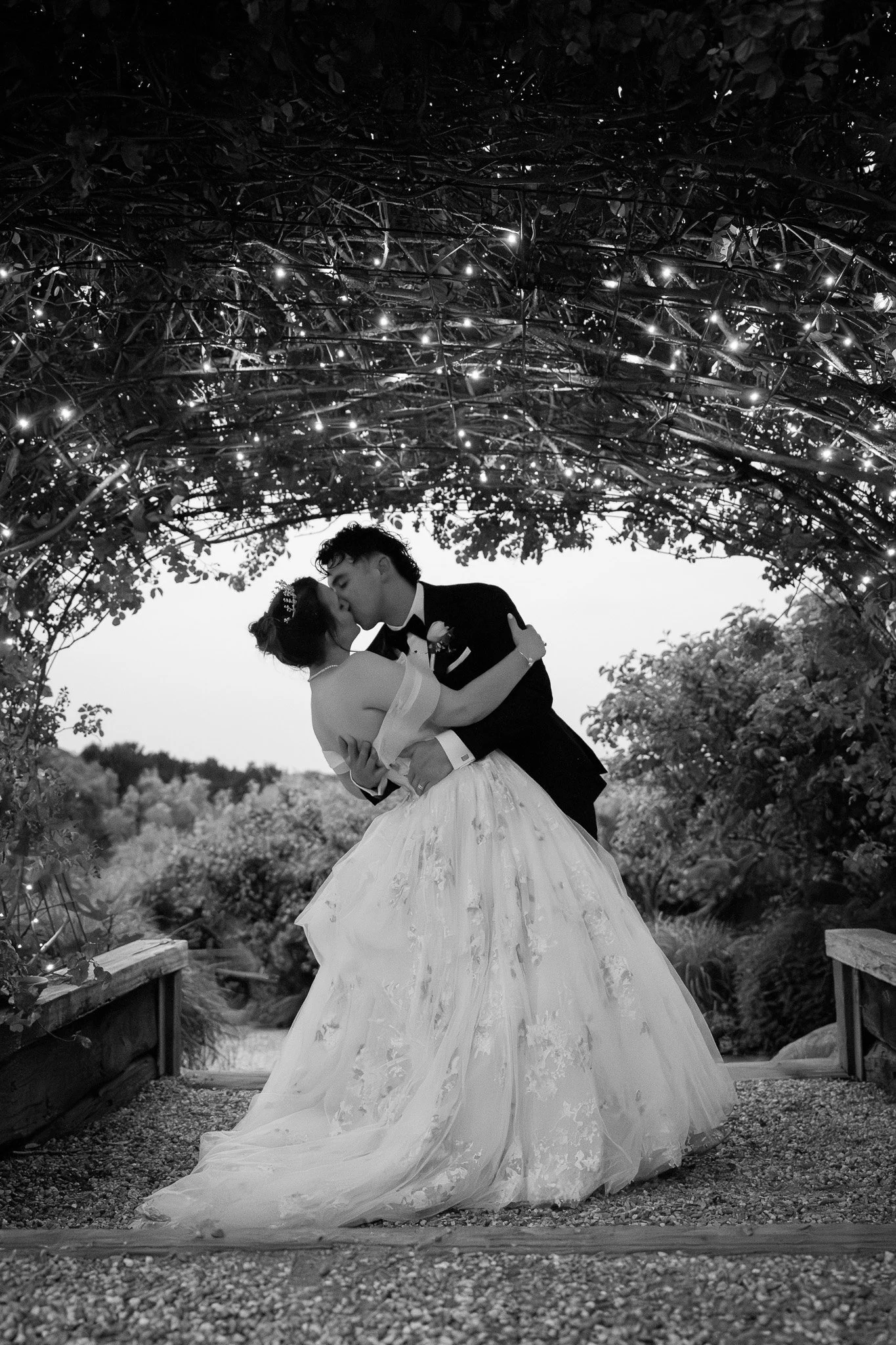 A black-and-white photo of a bride and groom sharing a kiss beneath an arch decorated with string lights, outdoors with trees in the background.