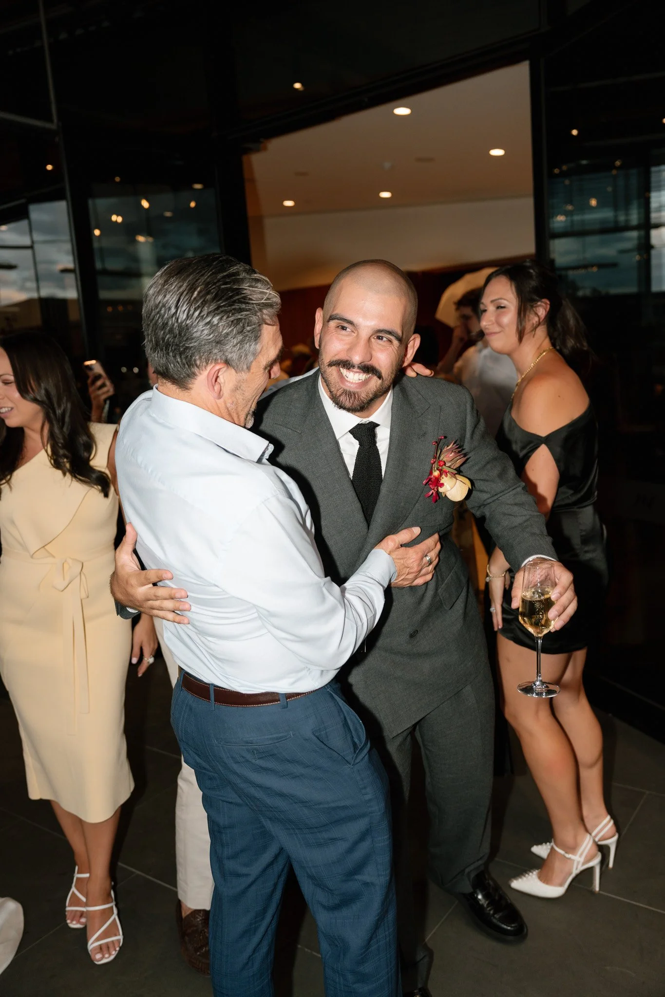 Two men are dancing and celebrating at a party, one holding a glass of champagne. They are smiling and enjoying their time, with other party guests in the background.