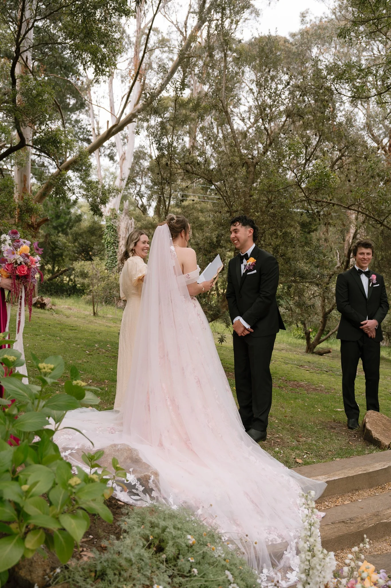 A couple getting married outdoors, with a bride in a pink wedding gown and a groom in a black tuxedo, standing in front of a officiant and an officiant's assistant. They are surrounded by trees and flowers.