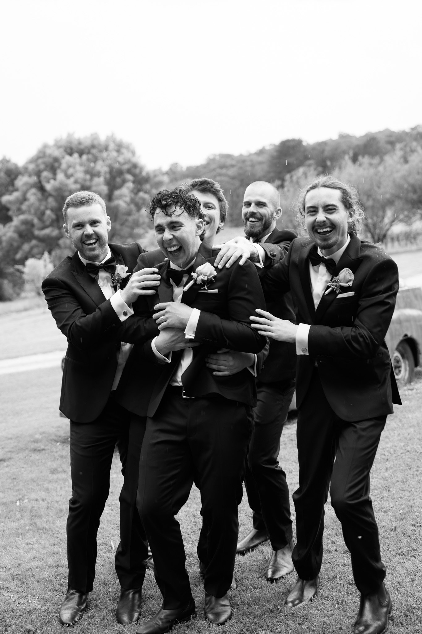 Group of five men in suits and bow ties laughing and hugging outdoors on a grassy field, with trees and cloudy sky in the background