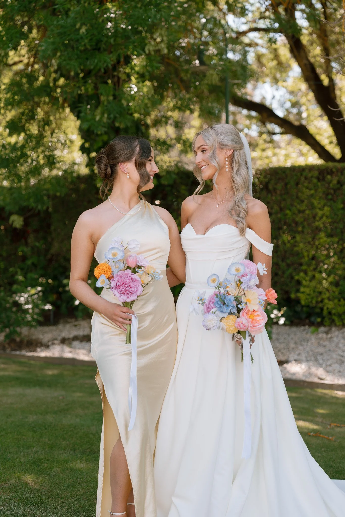Two women in wedding dresses holding colorful bouquets, standing outdoors on a lawn with trees and bushes in the background, smiling at each other.