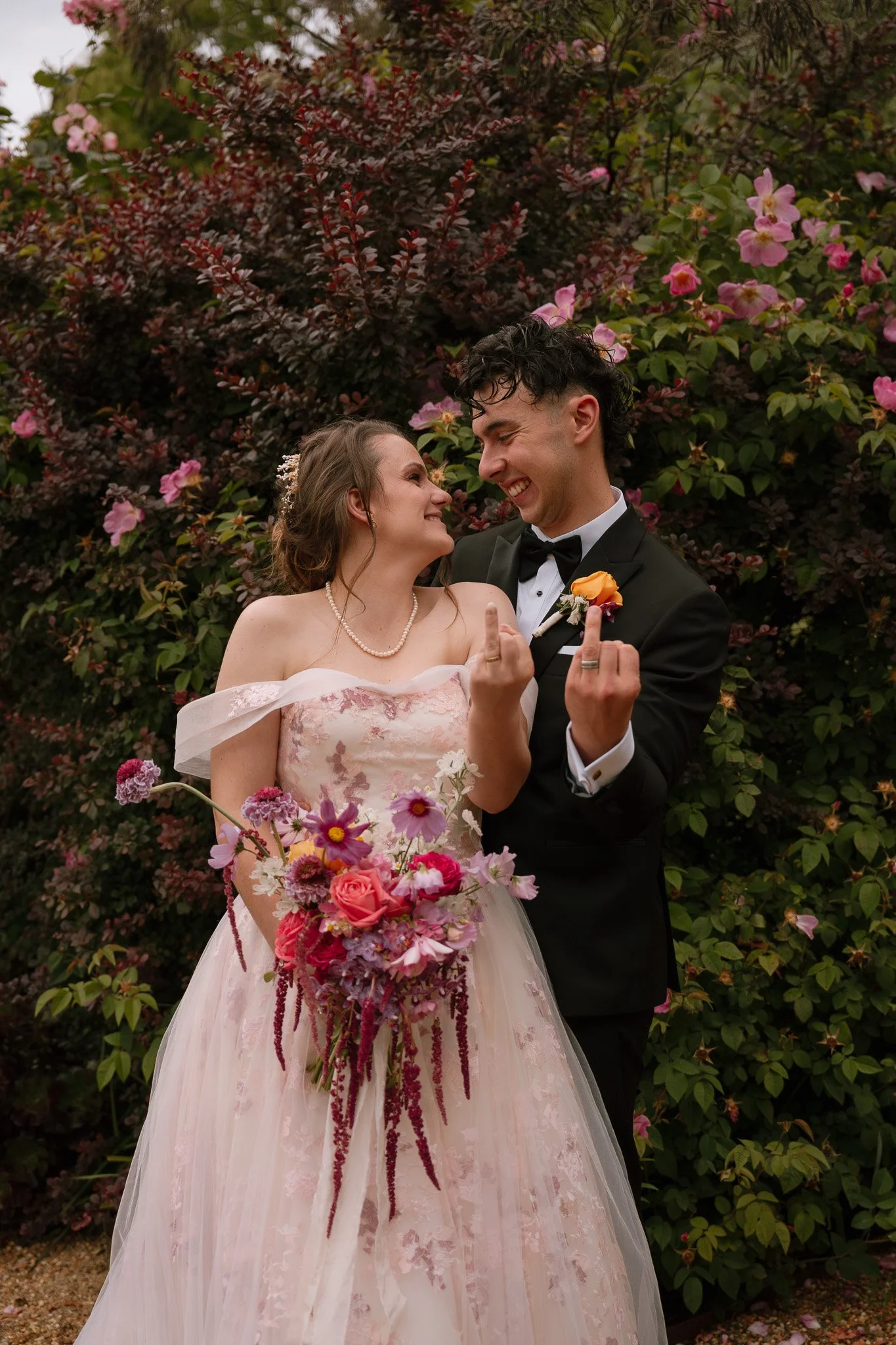 A bride and groom, standing close together outdoors, smiling and making rebellious gestures with their middle fingers, in front of blooming pink and purple flowers.