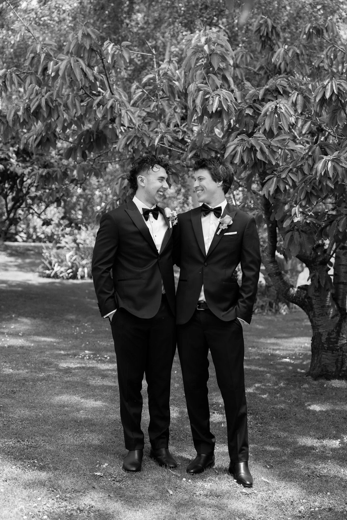 Two men in tuxedos with boutonnières, standing outdoors under a tree, smiling and looking at each other.