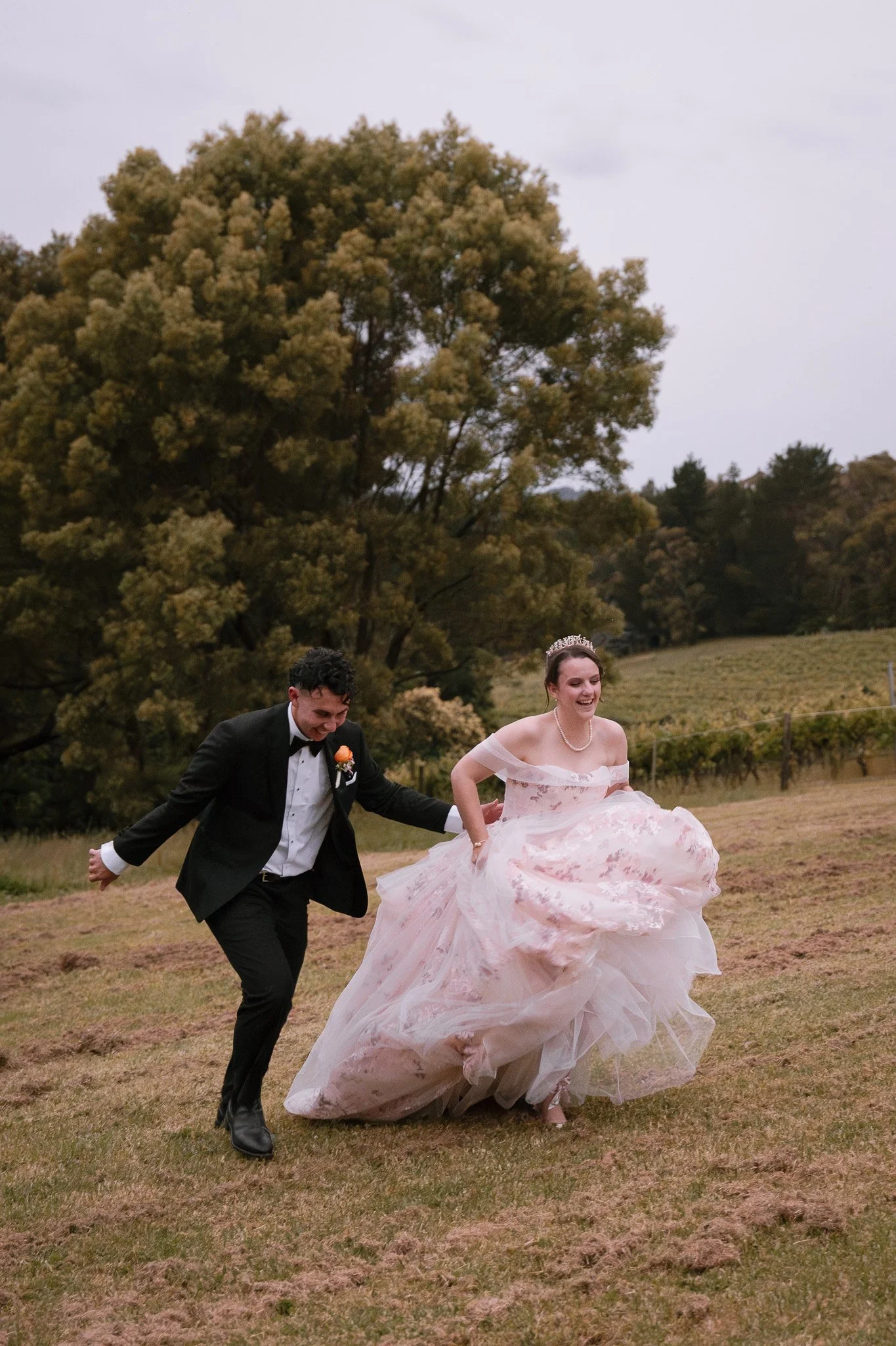 A bride and groom running outdoors on grass, smiling, with a large tree and hills in the background. The bride wears a pink and white gown with a tiara and pearl necklace. The groom wears a black tuxedo with a white shirt and bow tie.