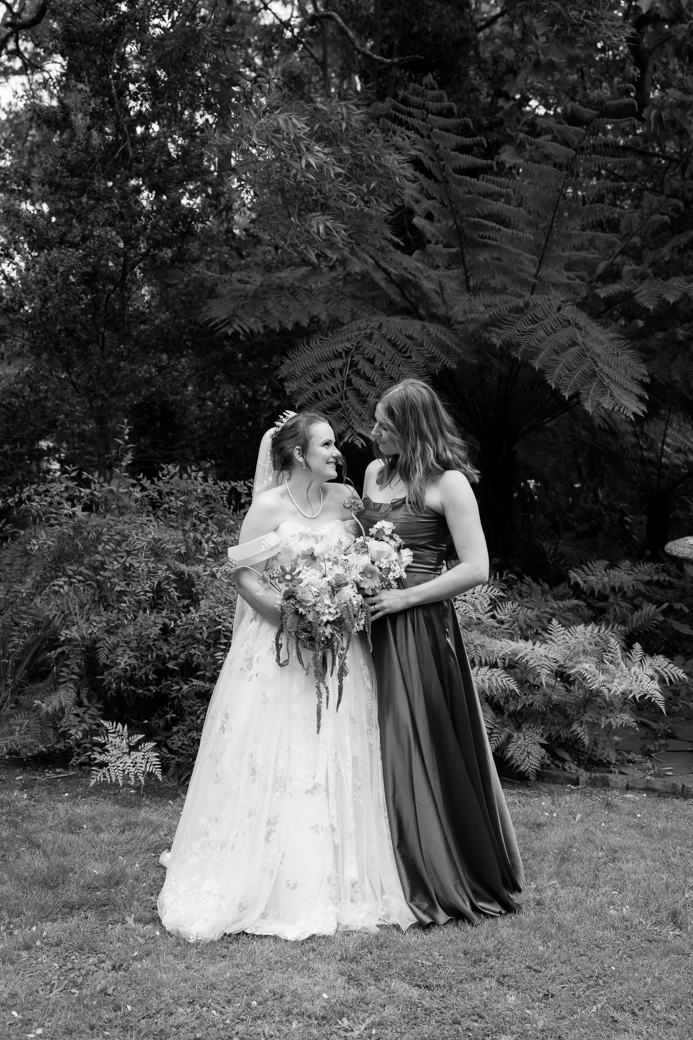 Black and white photograph of a bride in a wedding gown holding a bouquet, standing beside a woman in a dark formal dress, both smiling and looking at each other outdoors with lush foliage in the background.