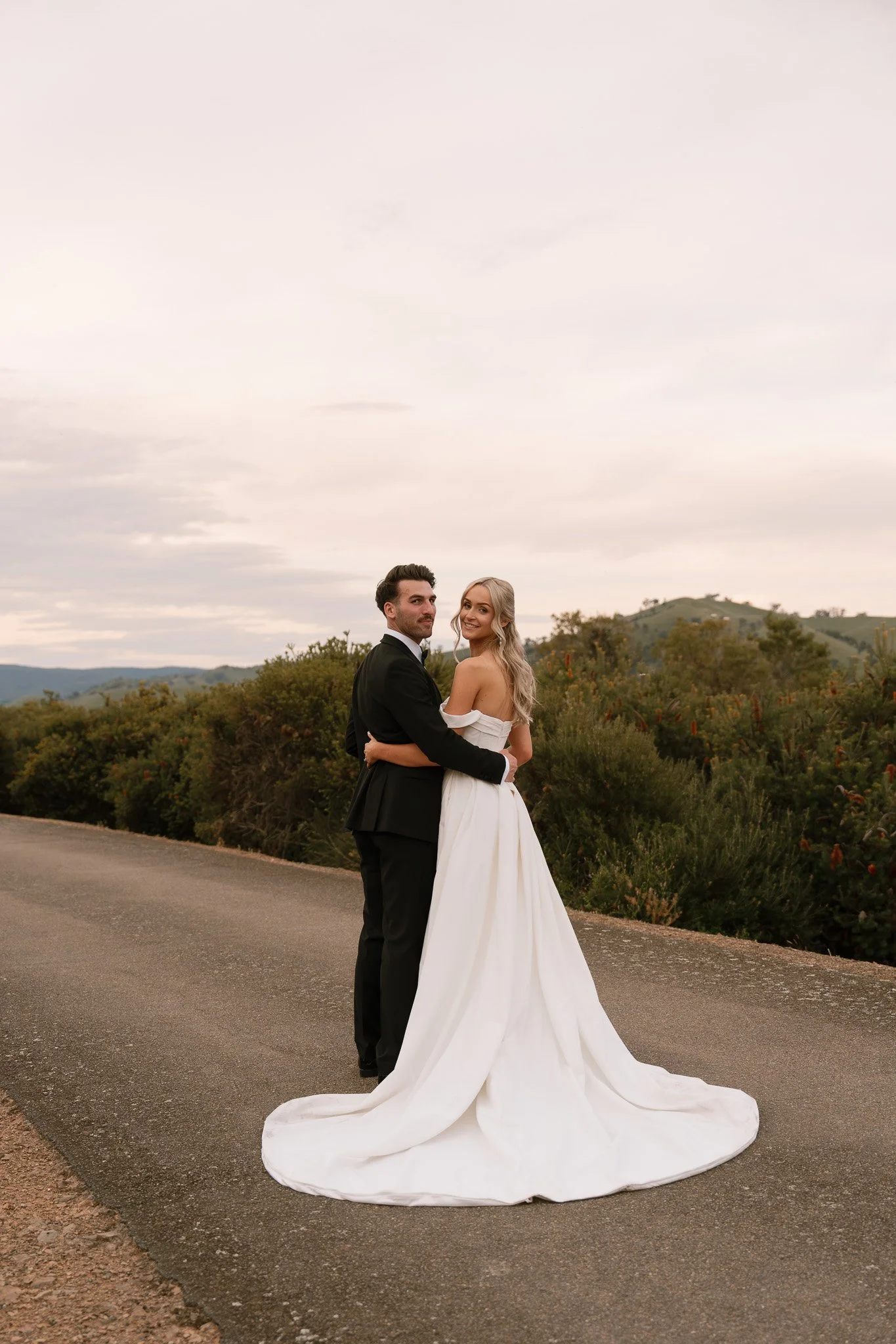 A bride and groom standing on a rural road, holding each other and smiling at the camera with a scenic hilly background under a cloudy sky.