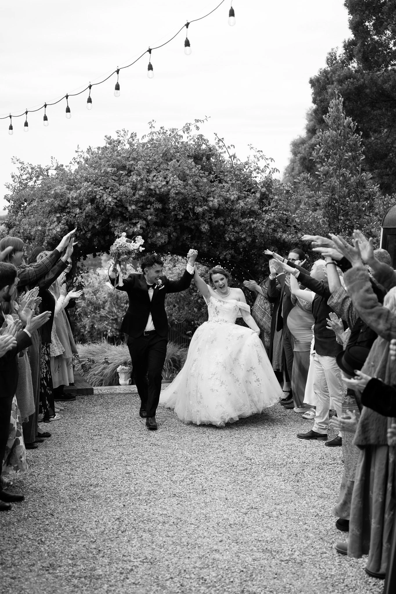 Black and white photo of a newlywed couple running and celebrating under a canopy of trees and string lights, surrounded by clapping guests. The groom is dressed in a suit, and the bride is wearing a wedding gown.