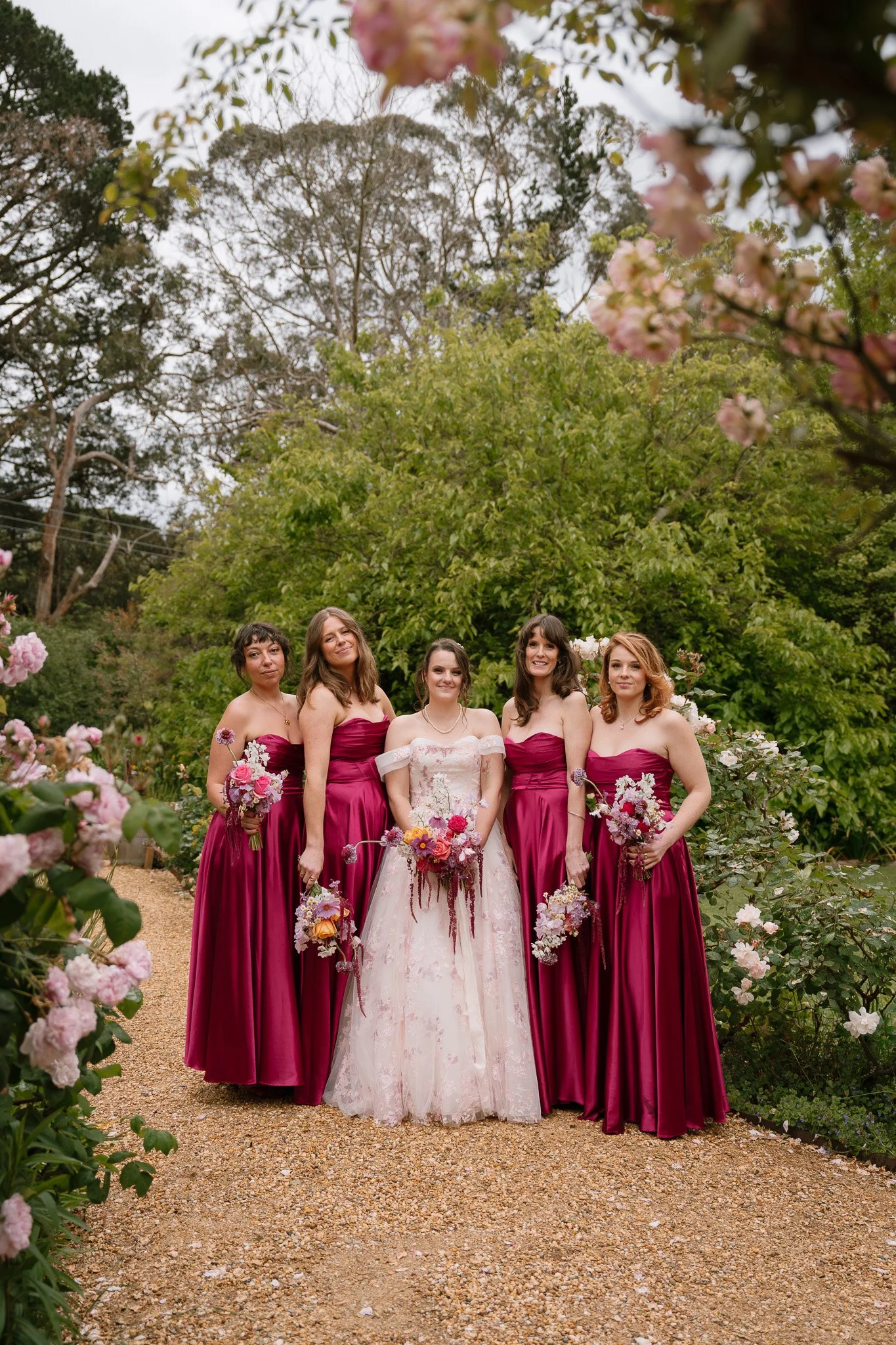 A bride in a white dress standing with four bridesmaids in magenta dresses holding bouquets, outdoors with trees and pink flowers.