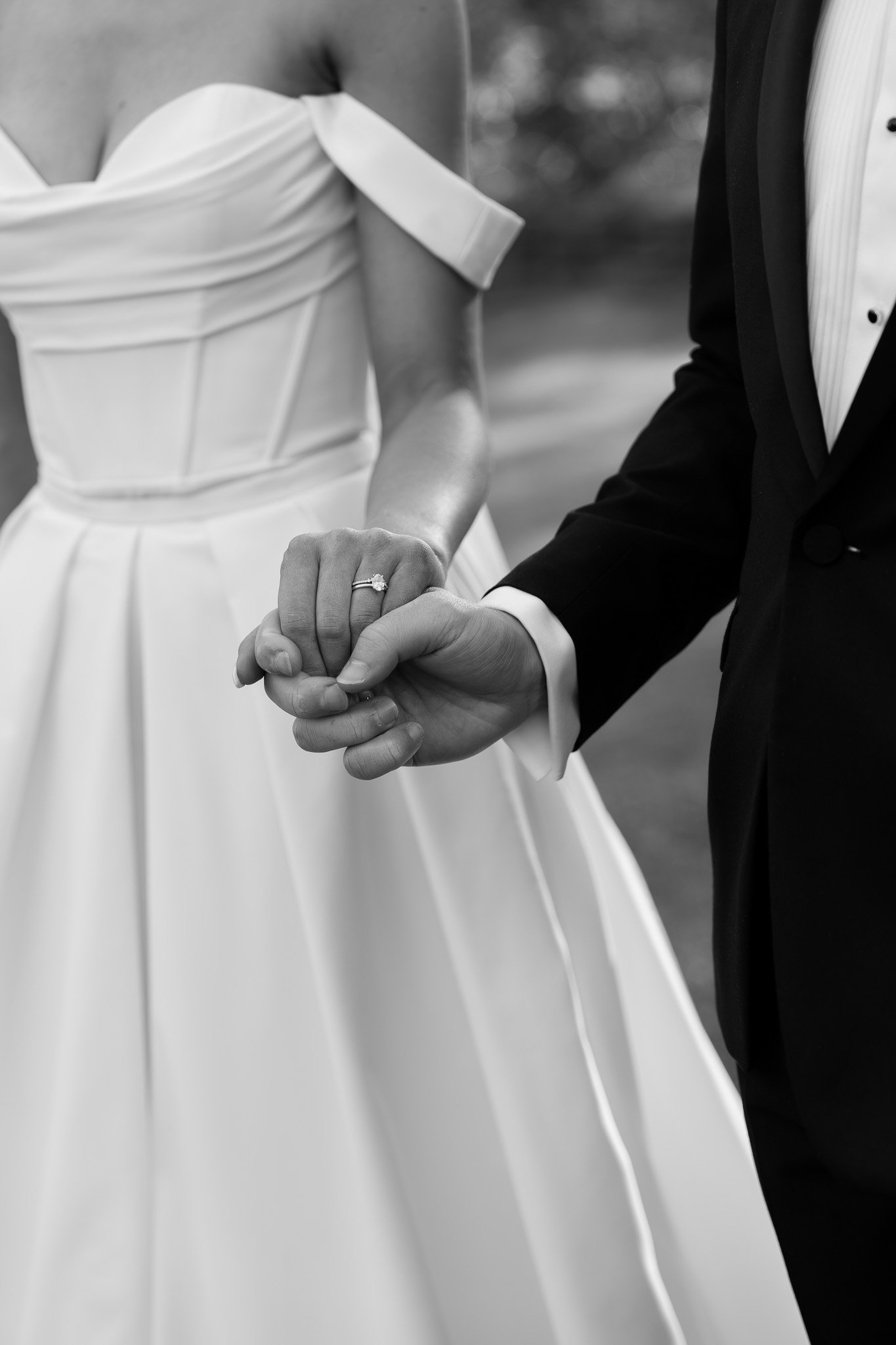 A bride and groom holding hands in a wedding scene, with the bride wearing a white dress and an engagement ring visible on her finger.