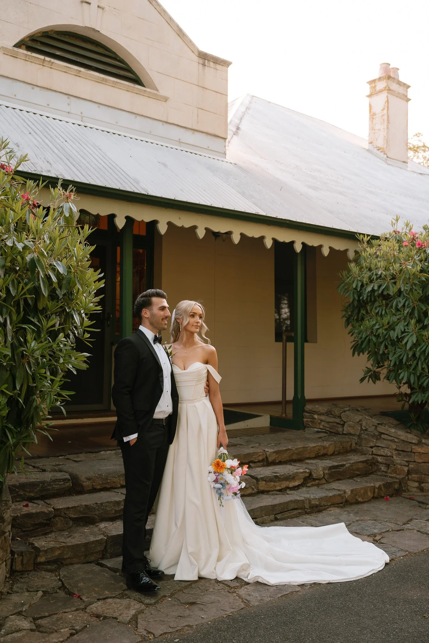 A bride and groom standing outside a house on their wedding day, with the bride holding a bouquet of flowers.